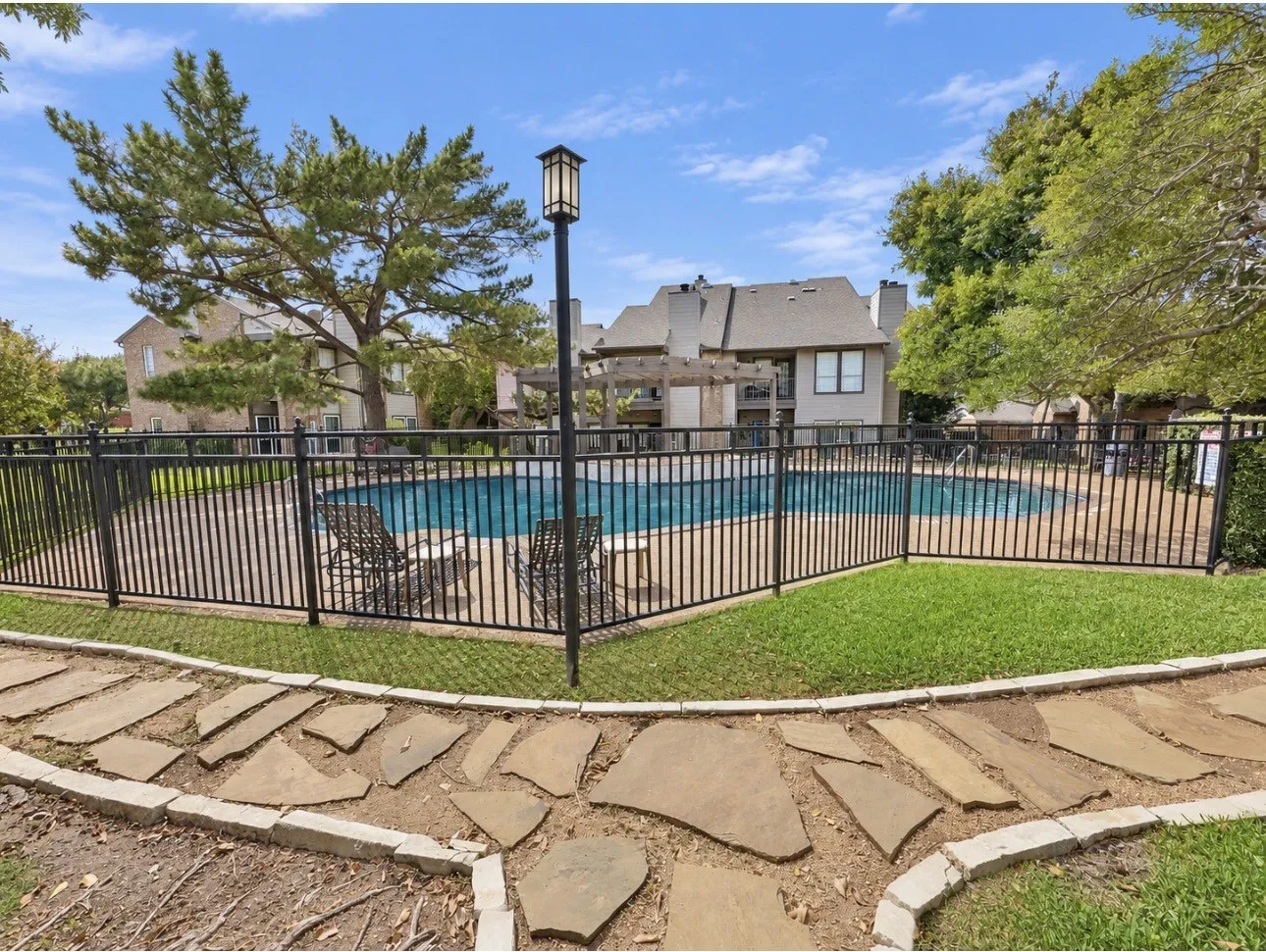 Paved flagstone pathway leading to a fenced swimming pool, surrounded by green grass and mature trees