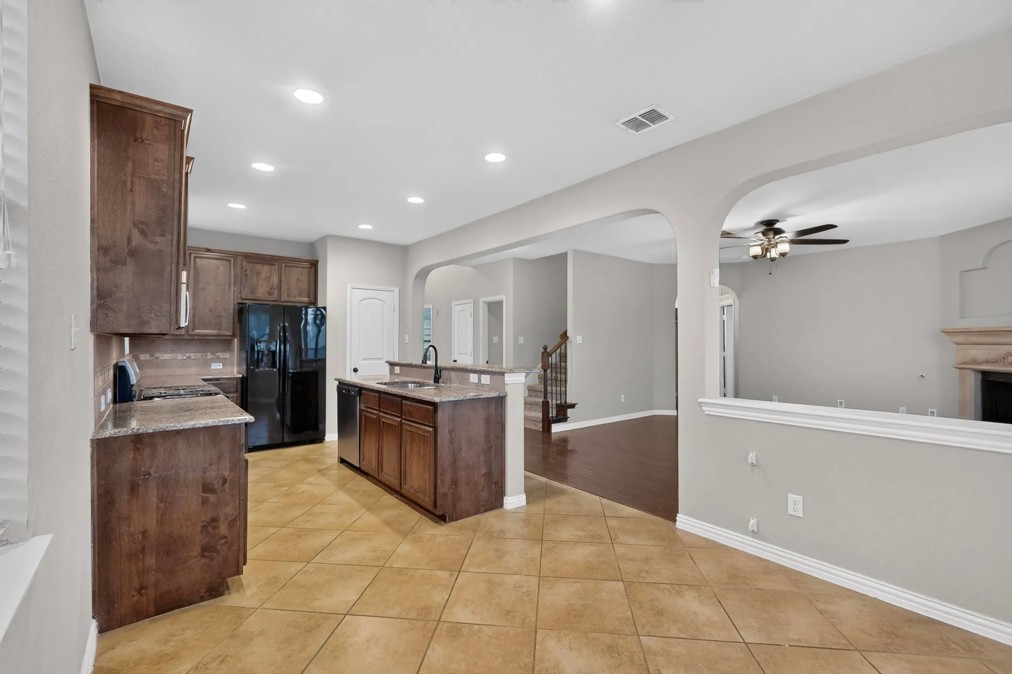 Spacious open-concept kitchen and living area featuring rich wood cabinetry, stainless steel appliances, a centered island with prep space, tile flooring, recessed lighting, and an arched passageway defining the adjoining​​‌​​​​‌​​‌‌​‌‌​​​‌‌​‌​‌​‌​​​‌​​ room