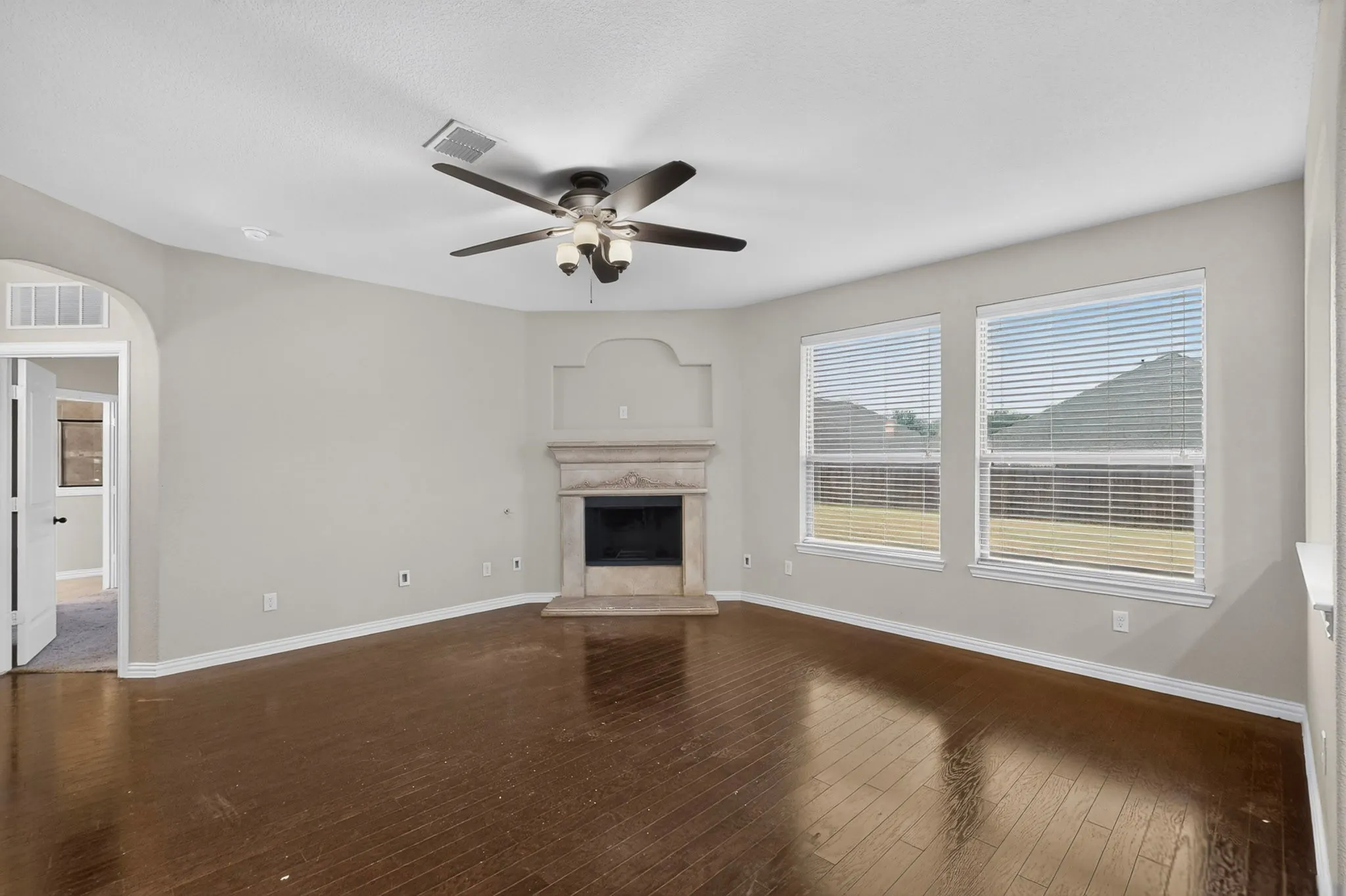 Hardwood-finish flooring, corner fireplace with stone surround, ceiling fan, and multiple windows with blinds