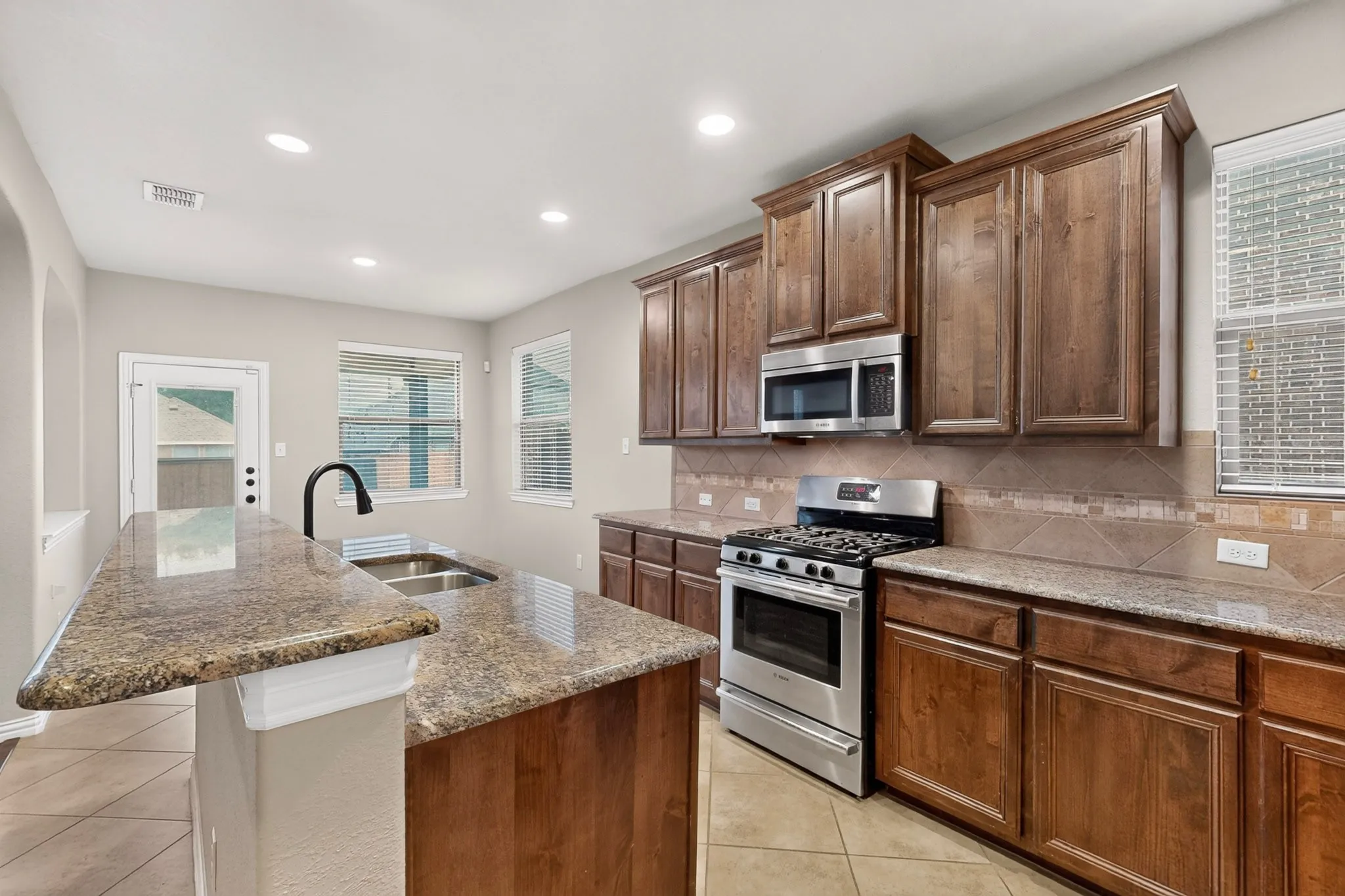 Spacious kitchen featuring an island with a double basin sink, stainless steel appliances, wood-finish cabinetry, and tiled flooring