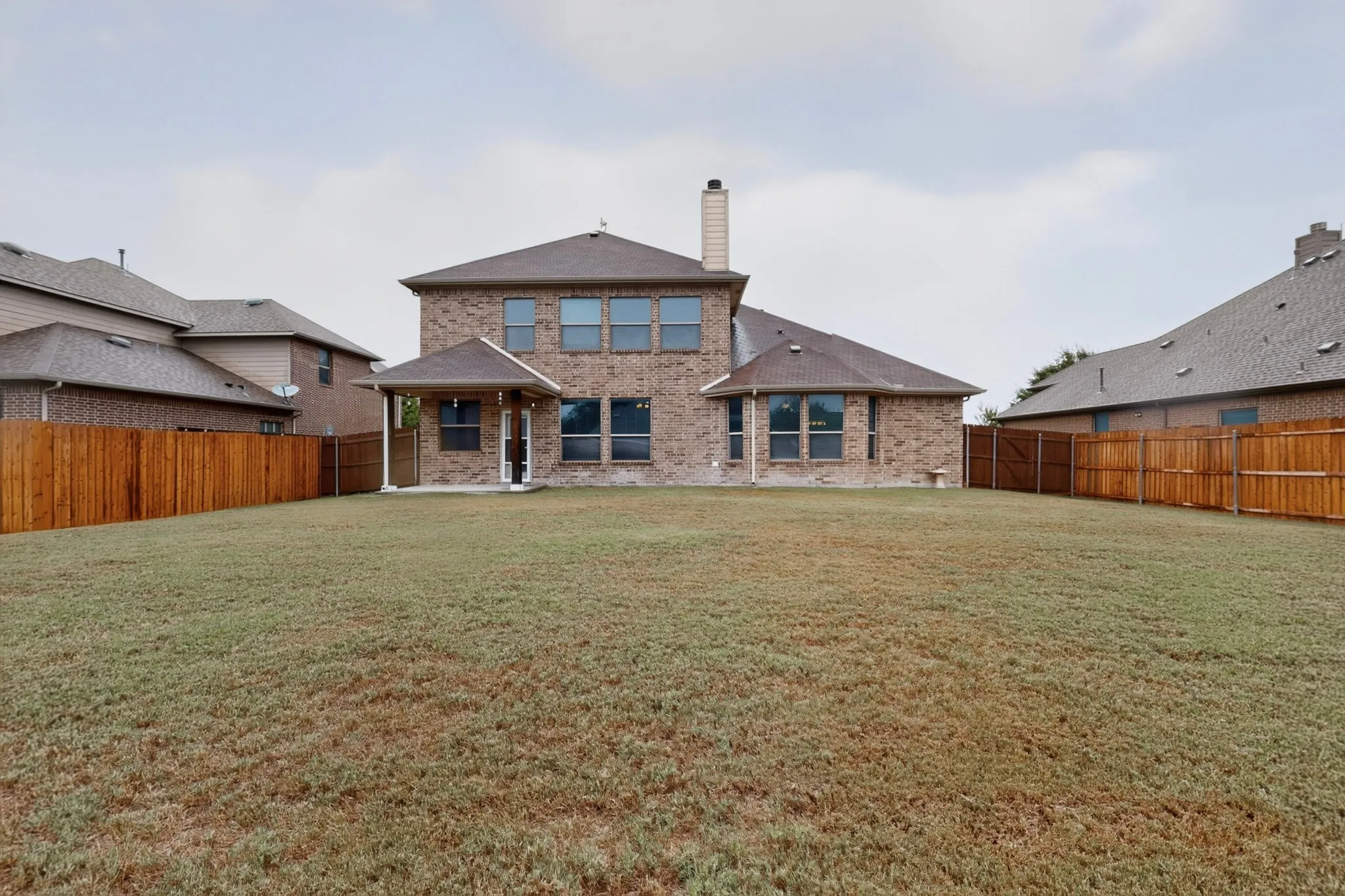 Expansive rear yard featuring a covered patio, brick exterior, multiple windows, and wood fencing