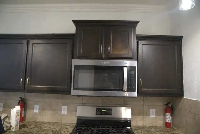 Dark wood-finish cabinetry, stainless steel microwave, stone-look countertop, tile backsplash, and crown molding