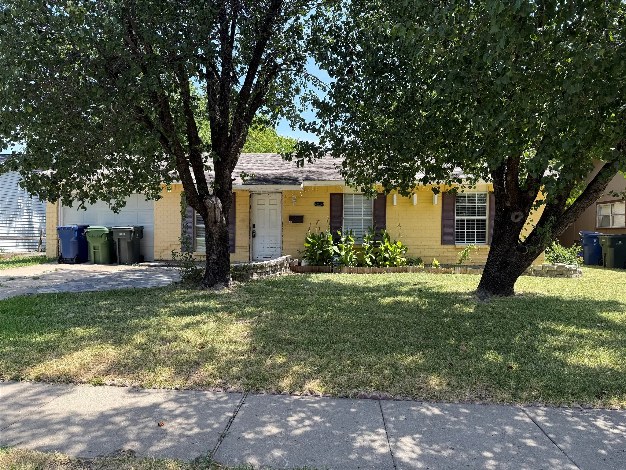 Property featuring a yellow brick exterior, multiple mature trees, a sidewalk, and a driveway