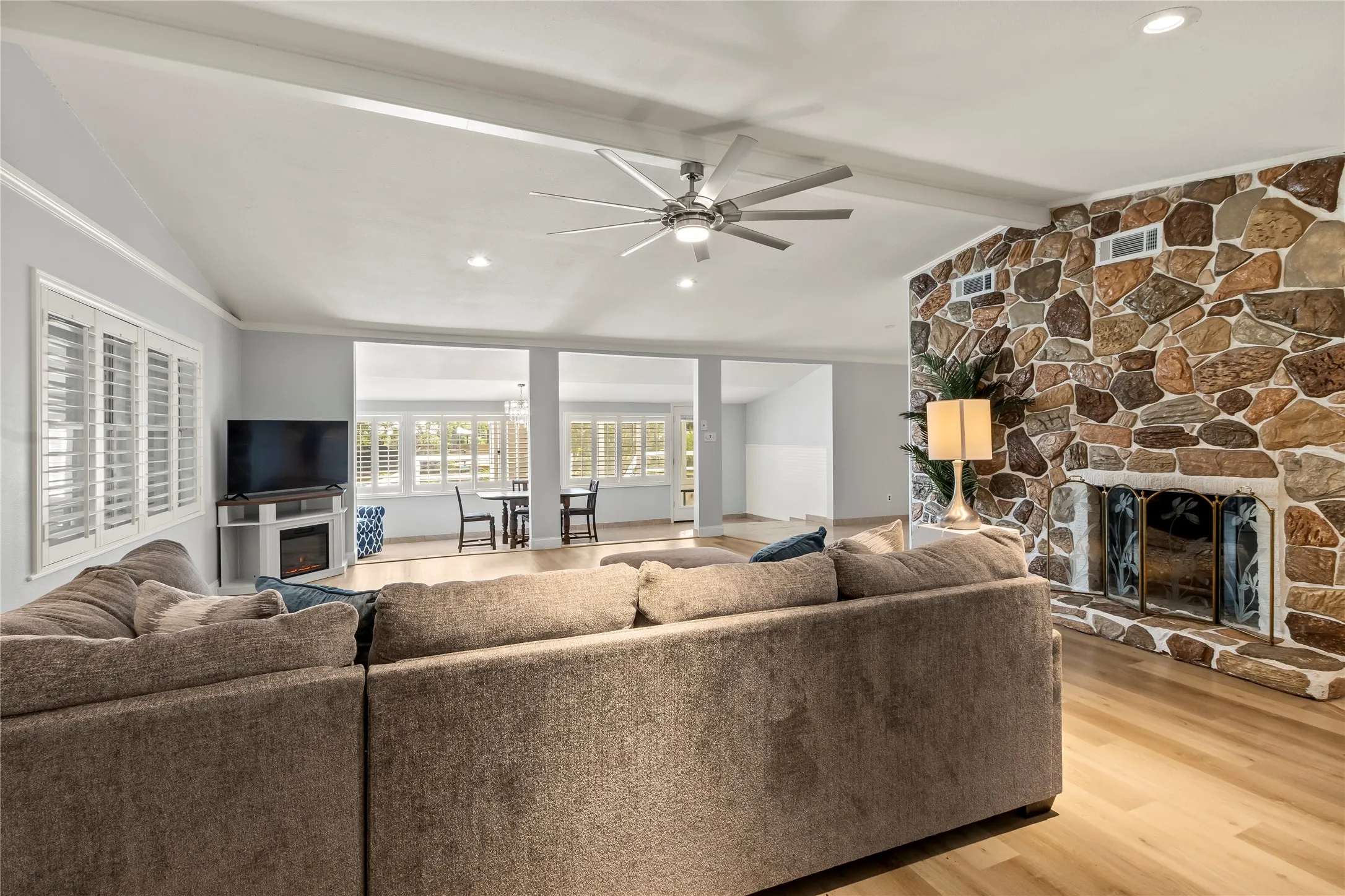 Vaulted ceiling with exposed beams, wood-finish flooring, stone-clad fireplace with hearth, recessed lighting, and white plantation shutters
