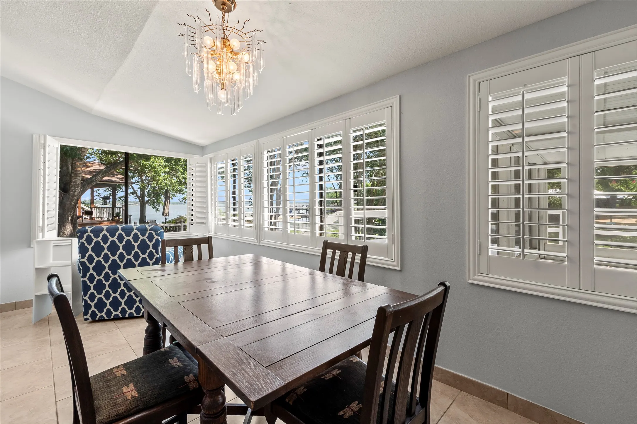 Tray ceiling with a decorative chandelier, multiple windows with white plantation shutters, and light-toned tile flooring