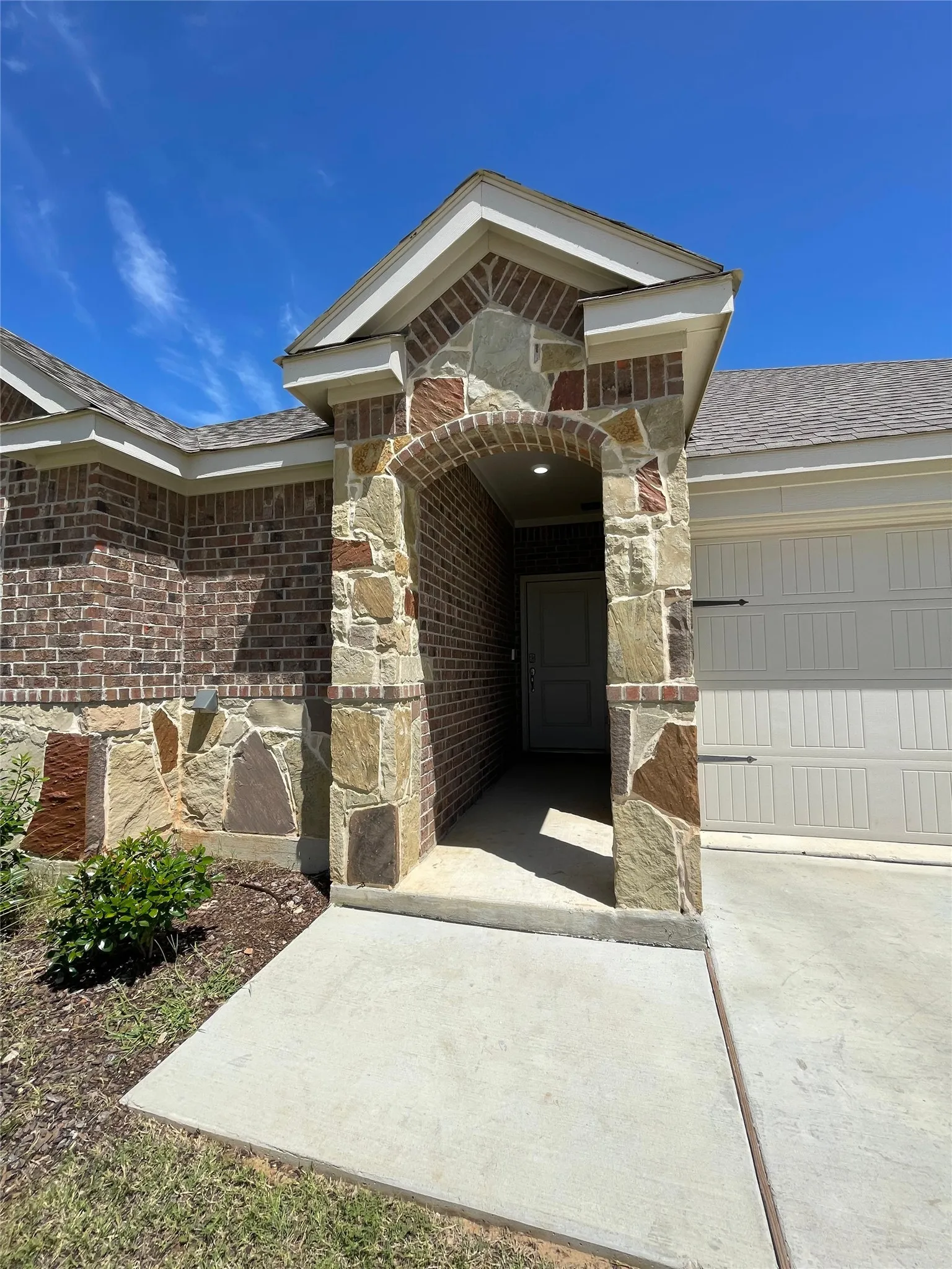 View of exterior entry with brick siding, stone siding, concrete driveway, a garage, and roof with shingles