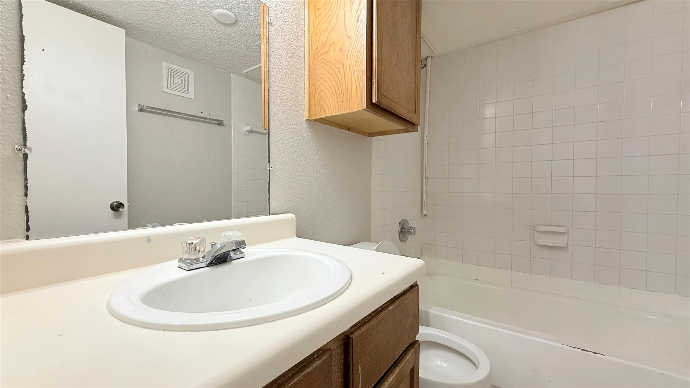 White tile tub surround, wood-finish vanity with integrated sink, and upper wood-finish cabinetry
