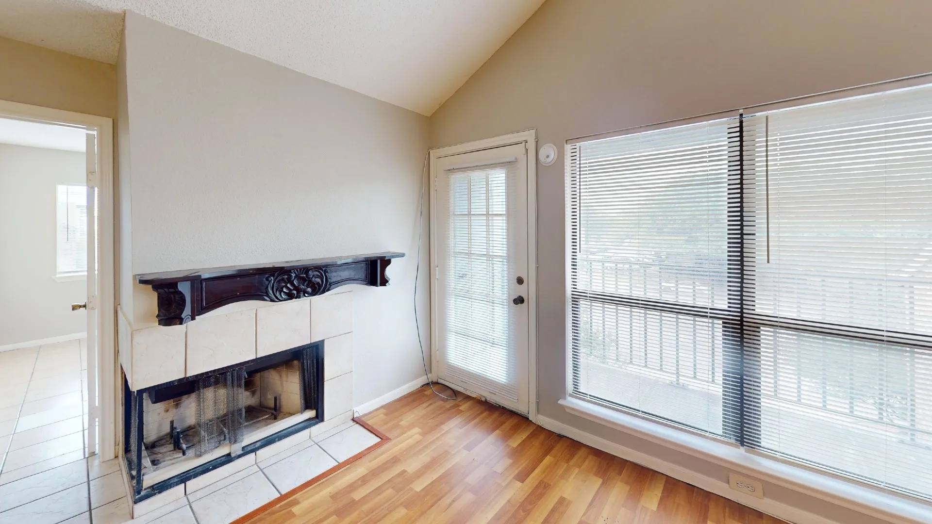Vaulted ceiling, wood-finish flooring, fireplace with tile surround and ornate mantel, glass-panel door, and large window with blinds