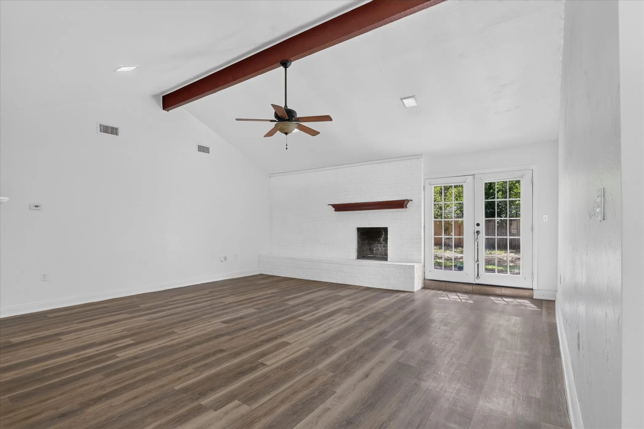 Vaulted ceilings with exposed beam, wood-finish flooring, white brick fireplace with mantel, ceiling fan, and double patio doors