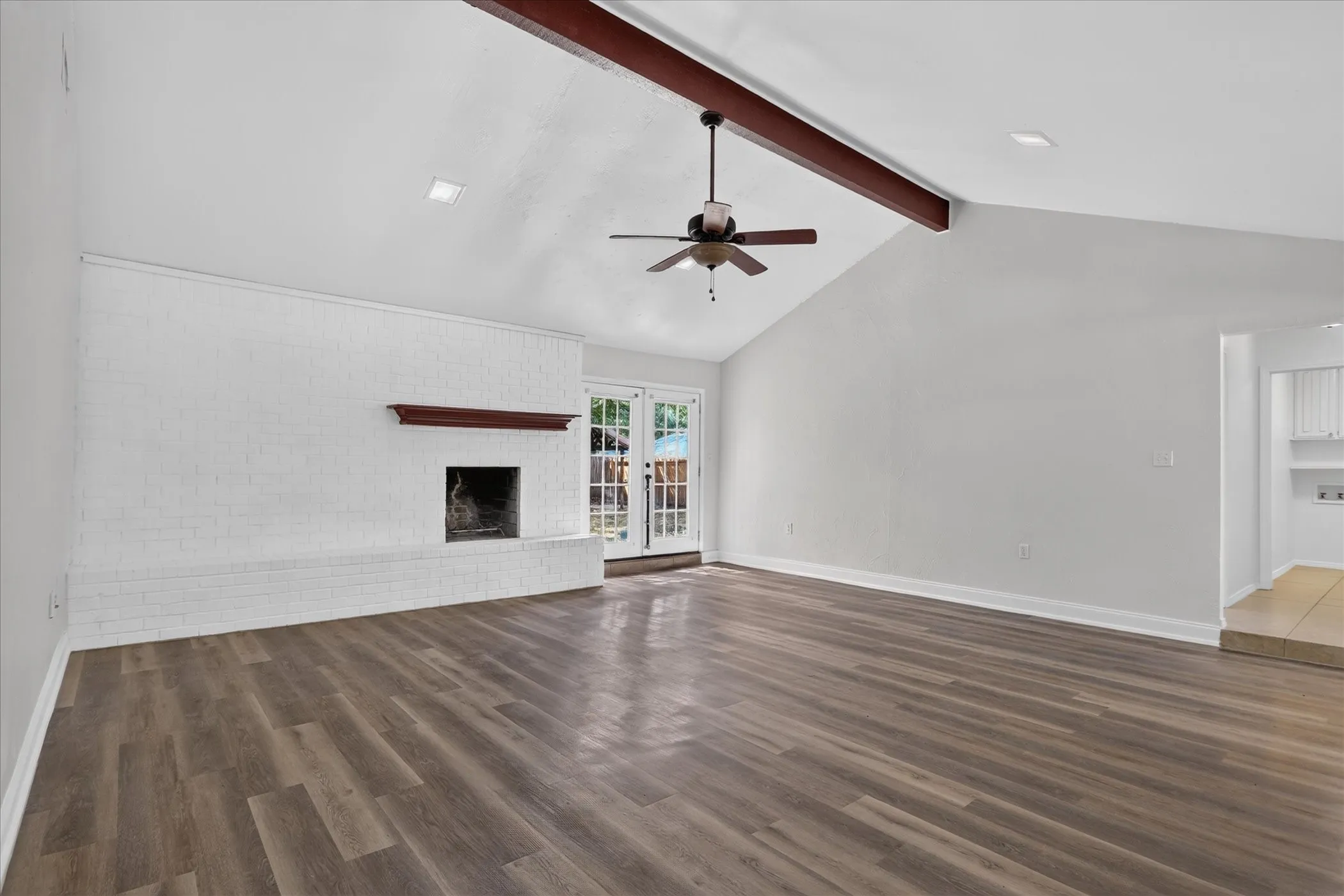 Vaulted ceiling with exposed beam, wood-finish flooring, white brick fireplace, ceiling fan, and French doors