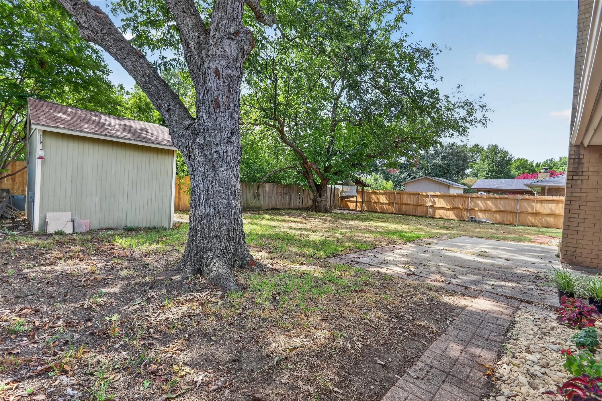 Expansive backyard featuring a mature tree, a detached storage shed, a wood fence, a concrete patio area, and a brick-finish pathway