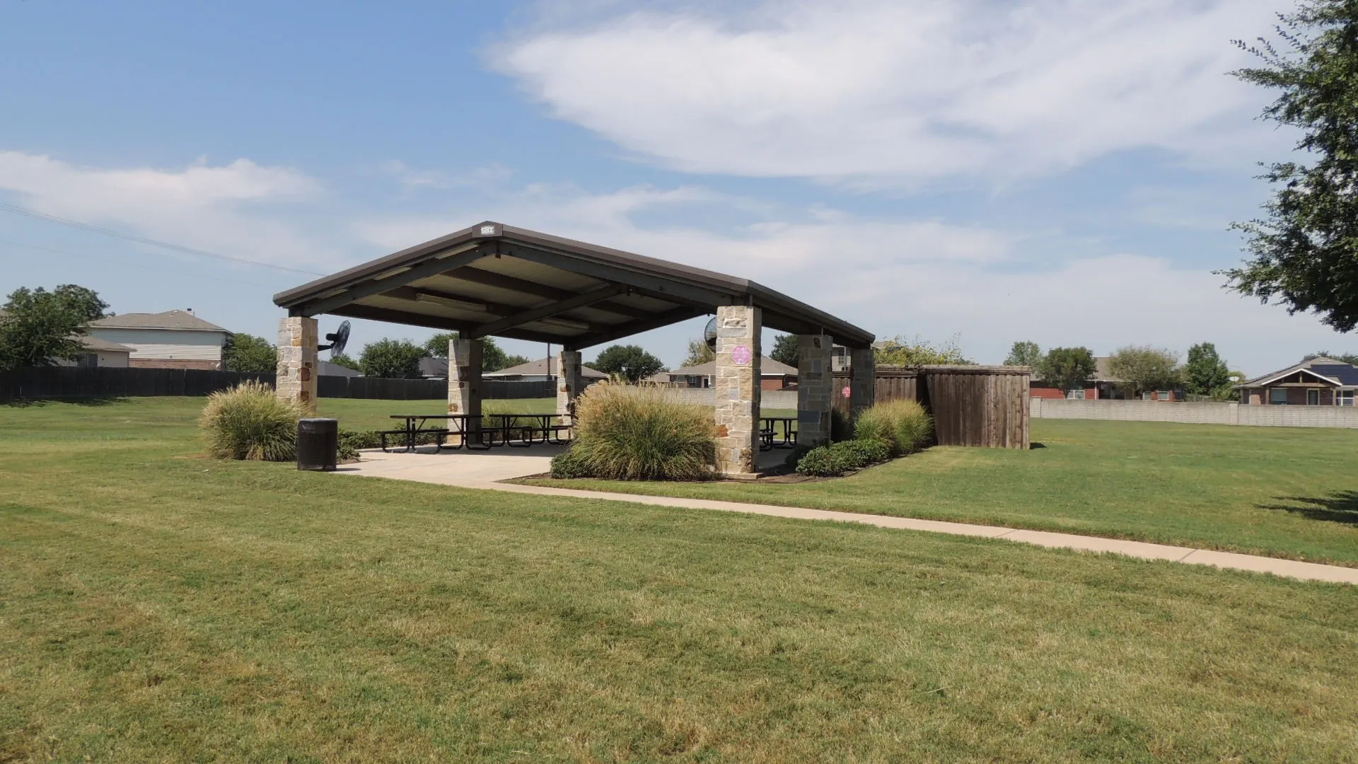 Covered outdoor pavilion with stone columns, a concrete pad, and dedicated walking paths, surrounded by expansive green space