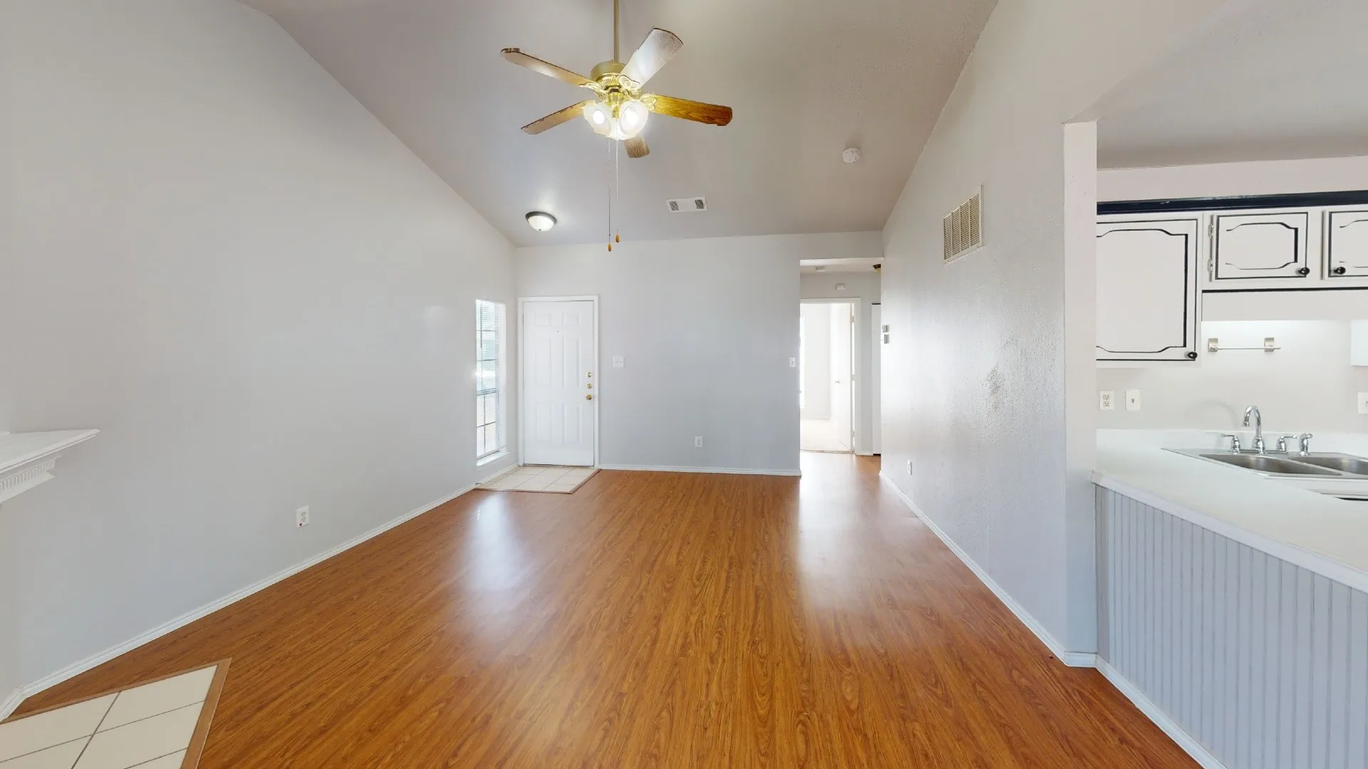 Vaulted ceiling, wood-finish flooring, ceiling fan, white paneled entry door, and a kitchen pass-through with beadboard paneling