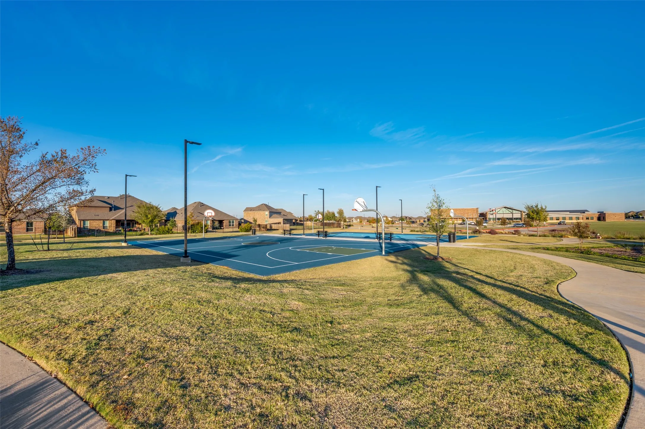 Recreational basketball court with multiple hoops, surrounded by landscaped green space and a paved walking path