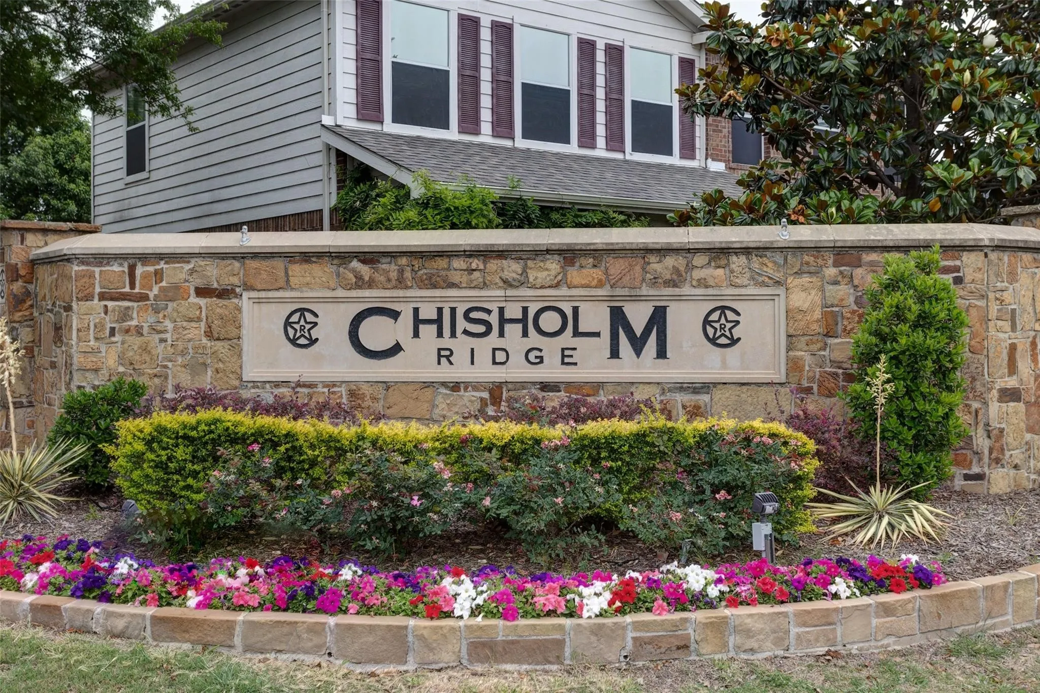 Stone-clad entry monument with a carved nameplate, featuring integrated landscaping with mature shrubs and colorful annuals