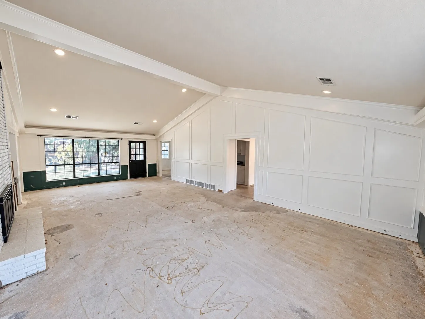 Vaulted ceiling with recessed lighting, large pane windows, white painted brick fireplace with black metal grate, paneled accent wall, and a black paneled door