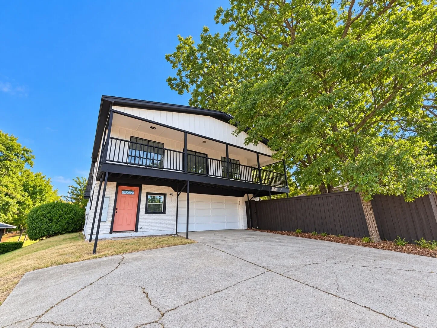 White exterior with black trim, a second-story balcony with black railings, a contrasting coral front door, an integrated garage, and a privacy fence