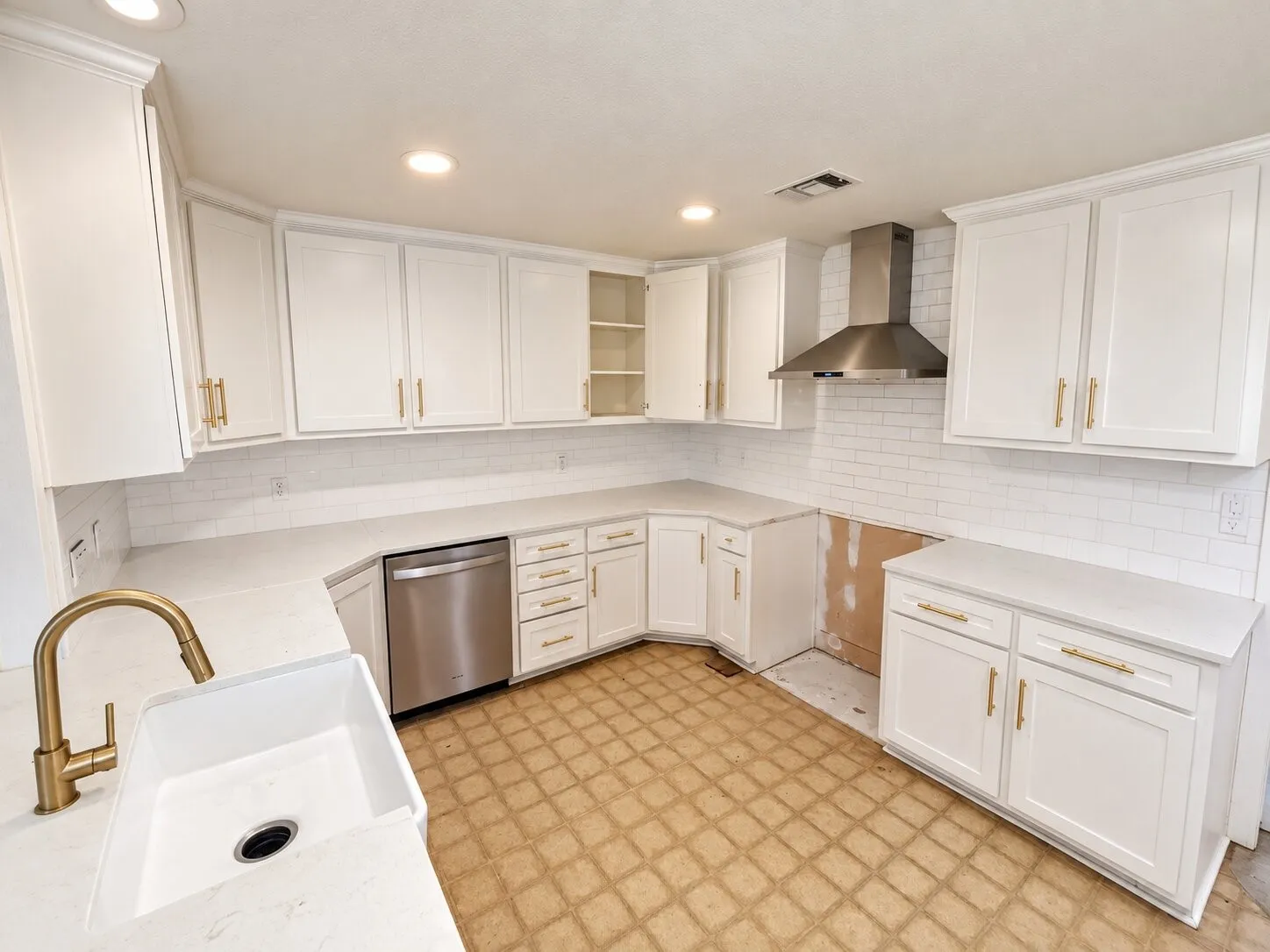 White cabinetry with gold-tone hardware, white subway tile backsplash, white countertop, farmhouse-style sink with gold-tone faucet, and a stainless steel range hood