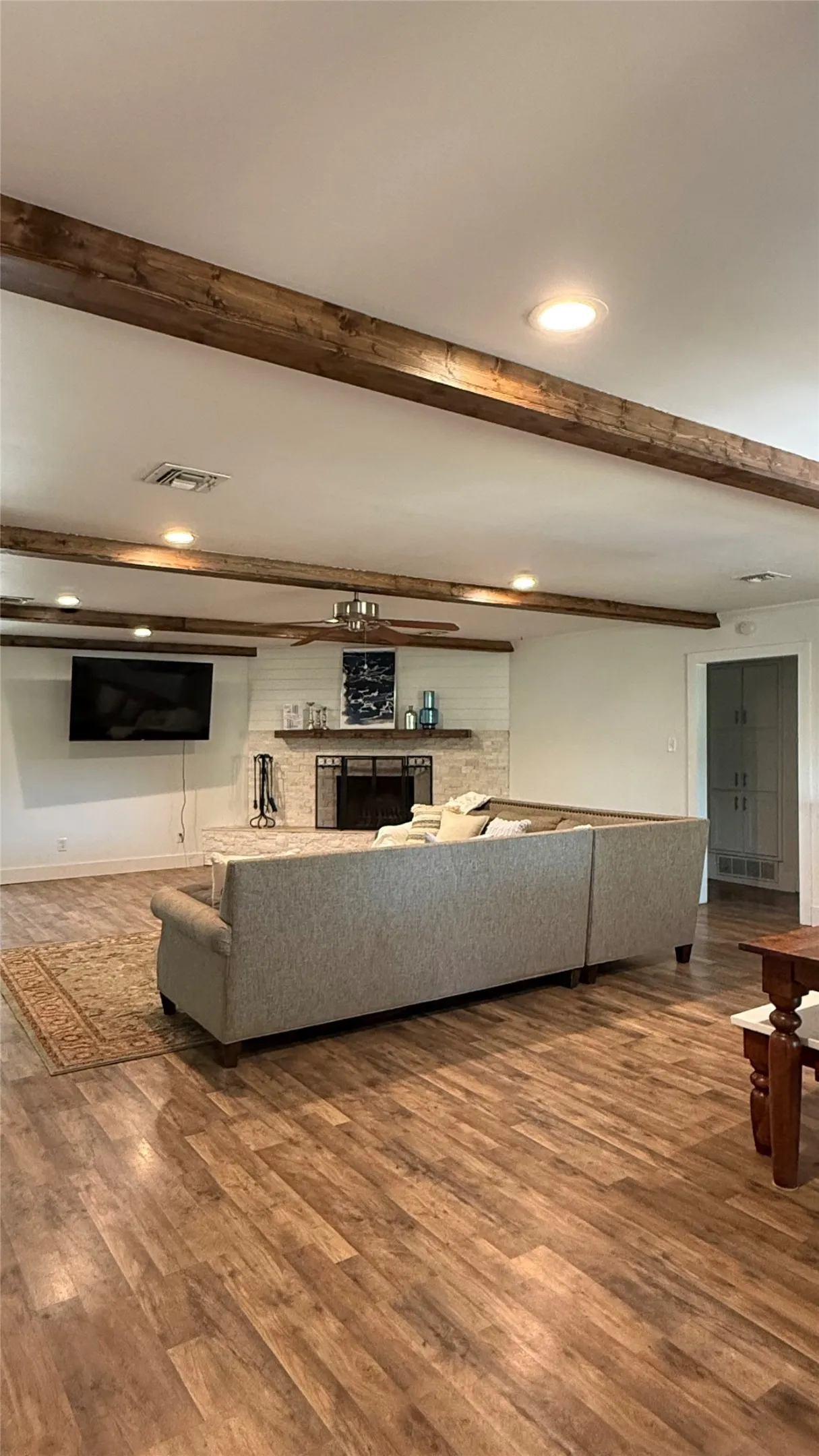 Wood-finish flooring throughout, ceiling with exposed wood beams and recessed lighting, fireplace with stone surround and shiplap accent wall, and a ceiling fan