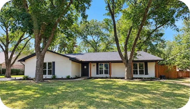White painted brick exterior with black window shutters, dark shingled roof, mature trees, and a wooden privacy fence