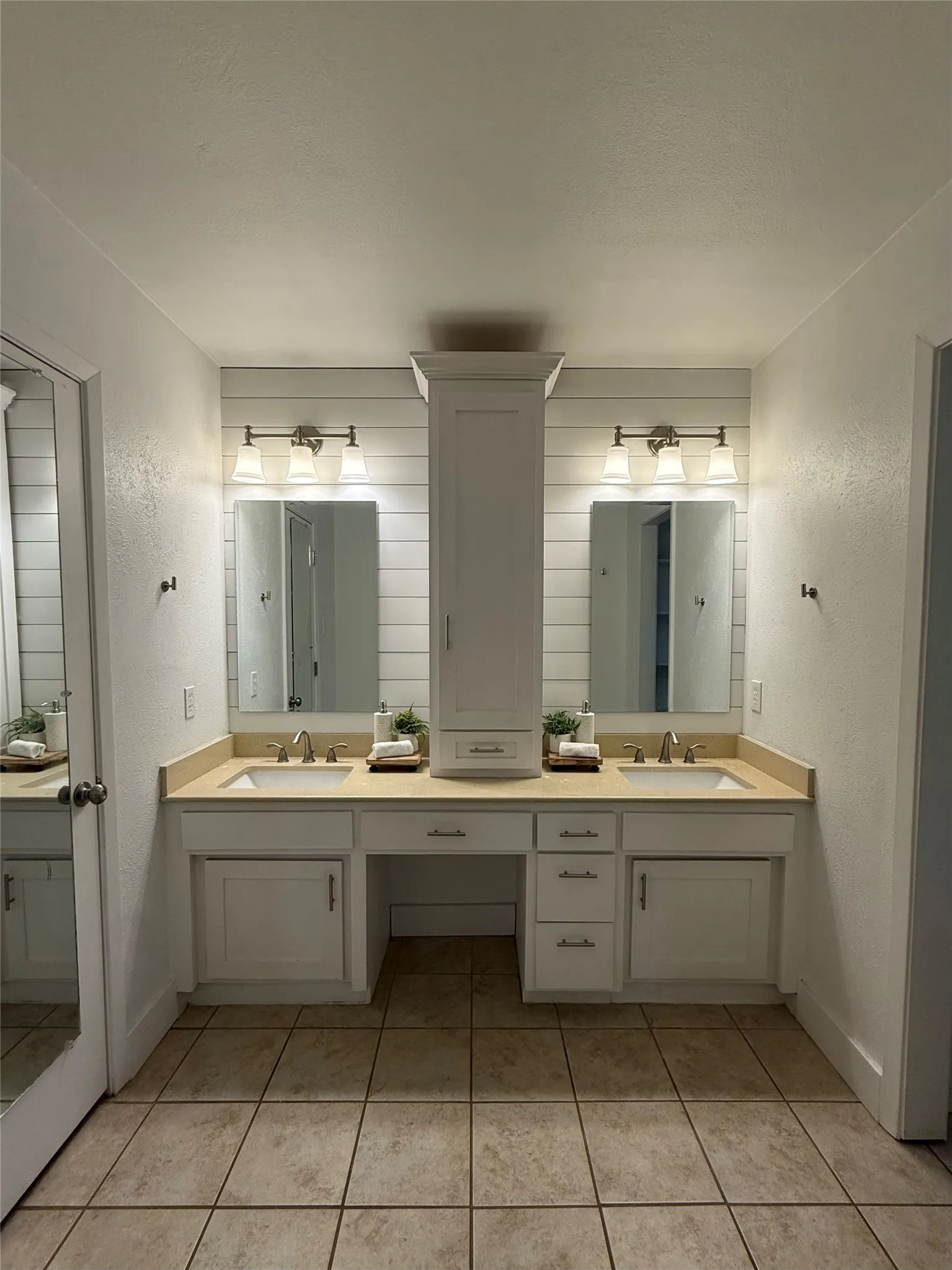 Dual vanity with sinks, light-toned countertops, white shaker-style cabinetry, continued shiplap and a central built-in storage tower. The closet is to the left and separate shower and toilet are the the right.