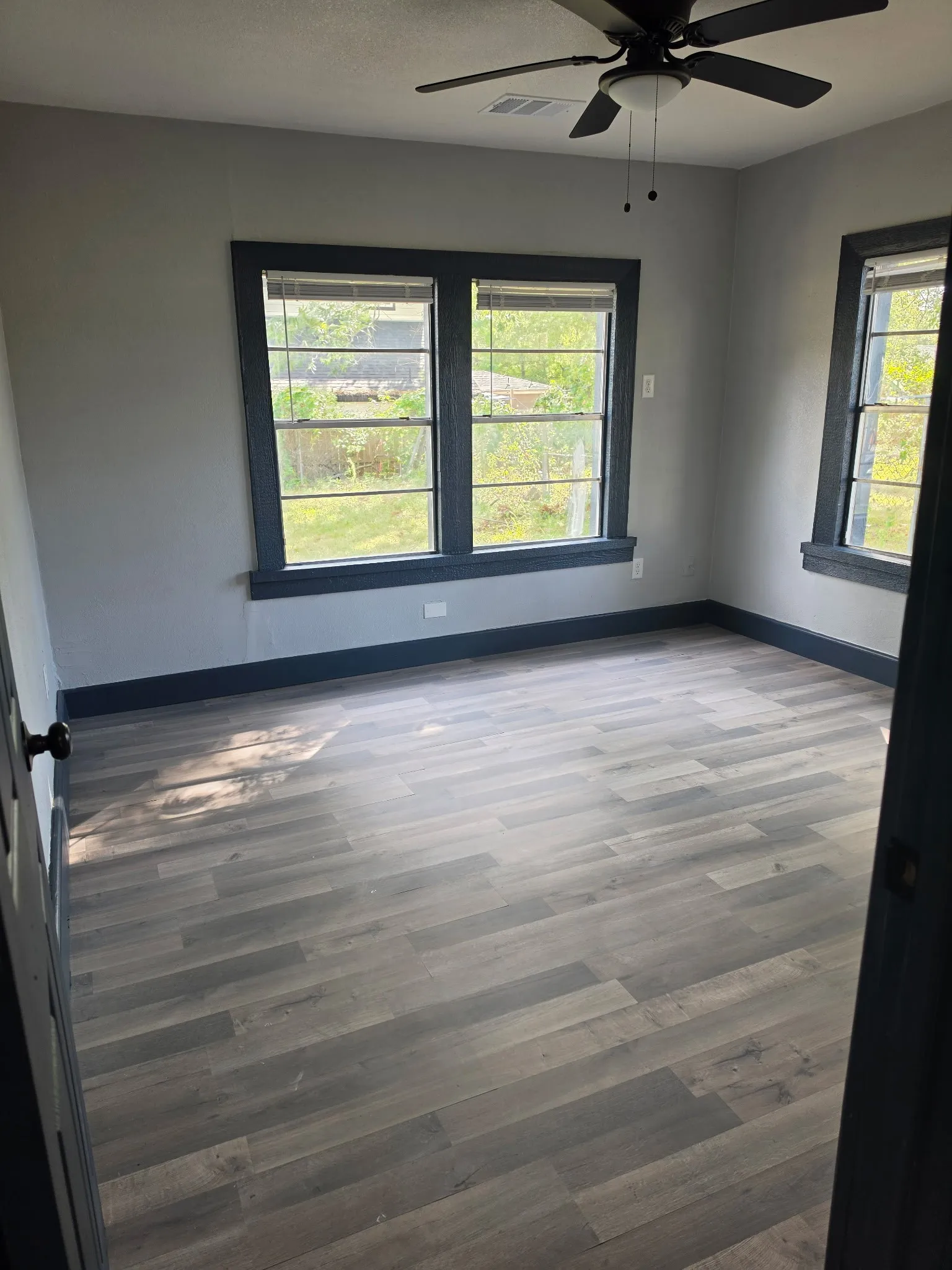 Spacious room featuring wood-finish flooring, multiple windows, a ceiling fan with light fixture, and dark trim details