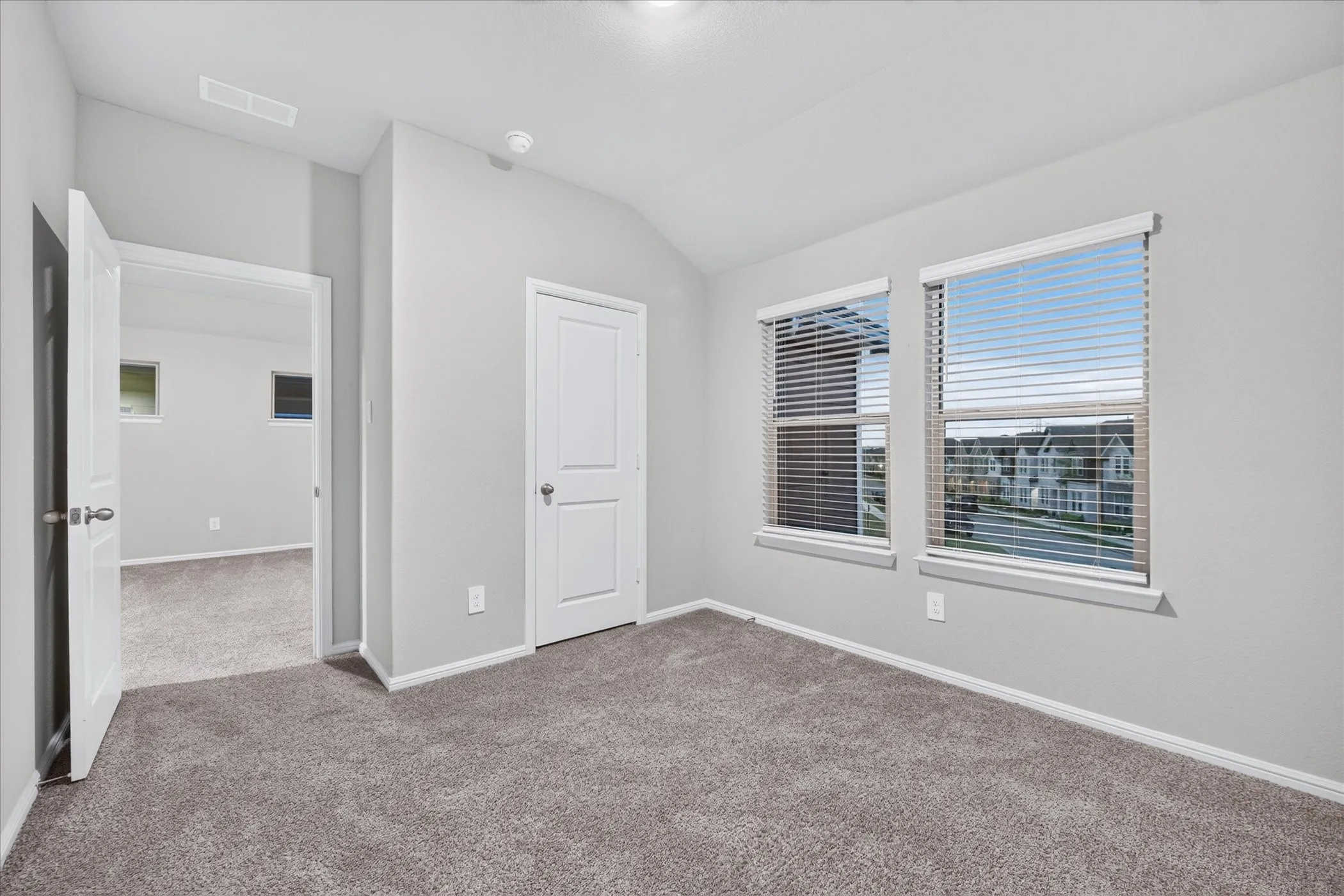 Carpeted room featuring two windows with blinds, a white paneled door, and a vaulted ceiling