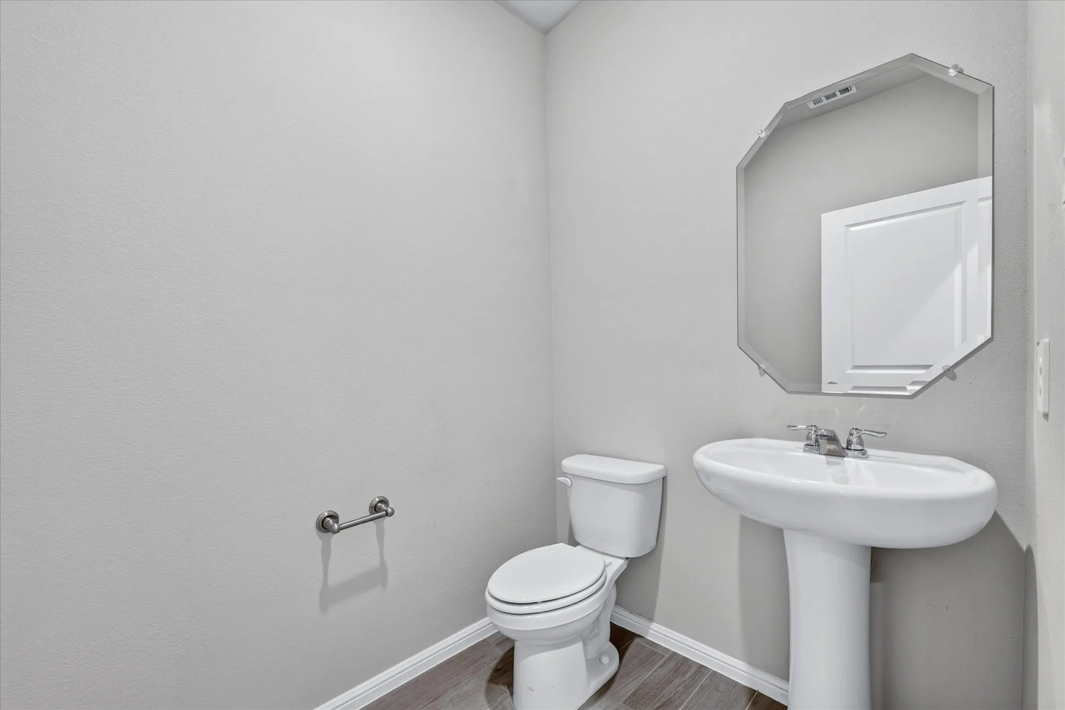 Pedestal sink with chrome fixtures, octagonal frameless mirror, and wood-finish flooring