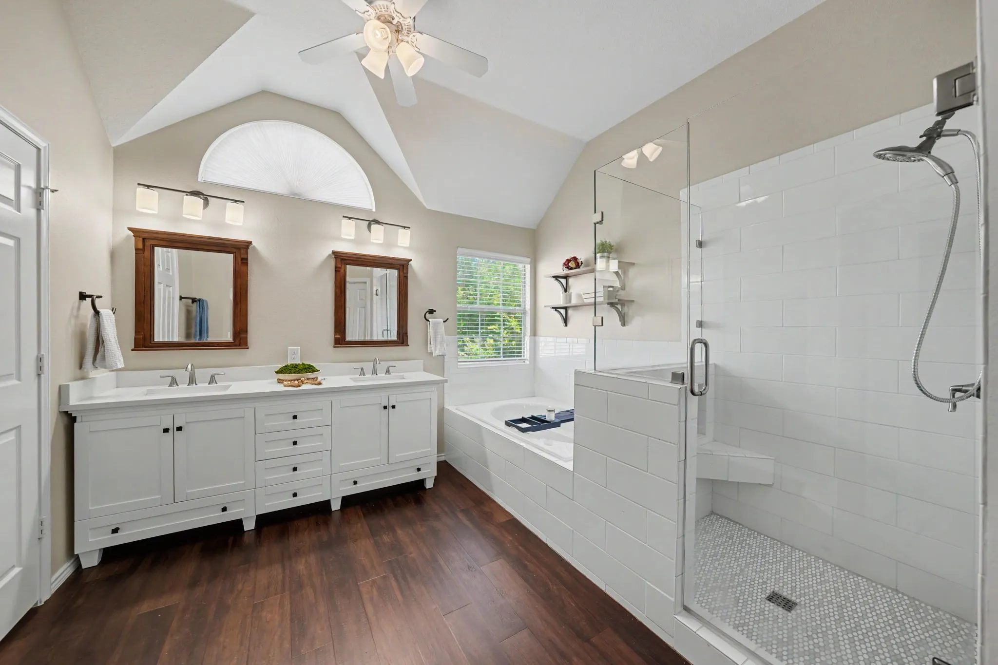 Vaulted ceiling with an arched window, dual vanity with white cabinetry and integrated sinks, walk-in shower with glass enclosure, white subway tile surround, and mosaic tile flooring, and a built-in soaking tub