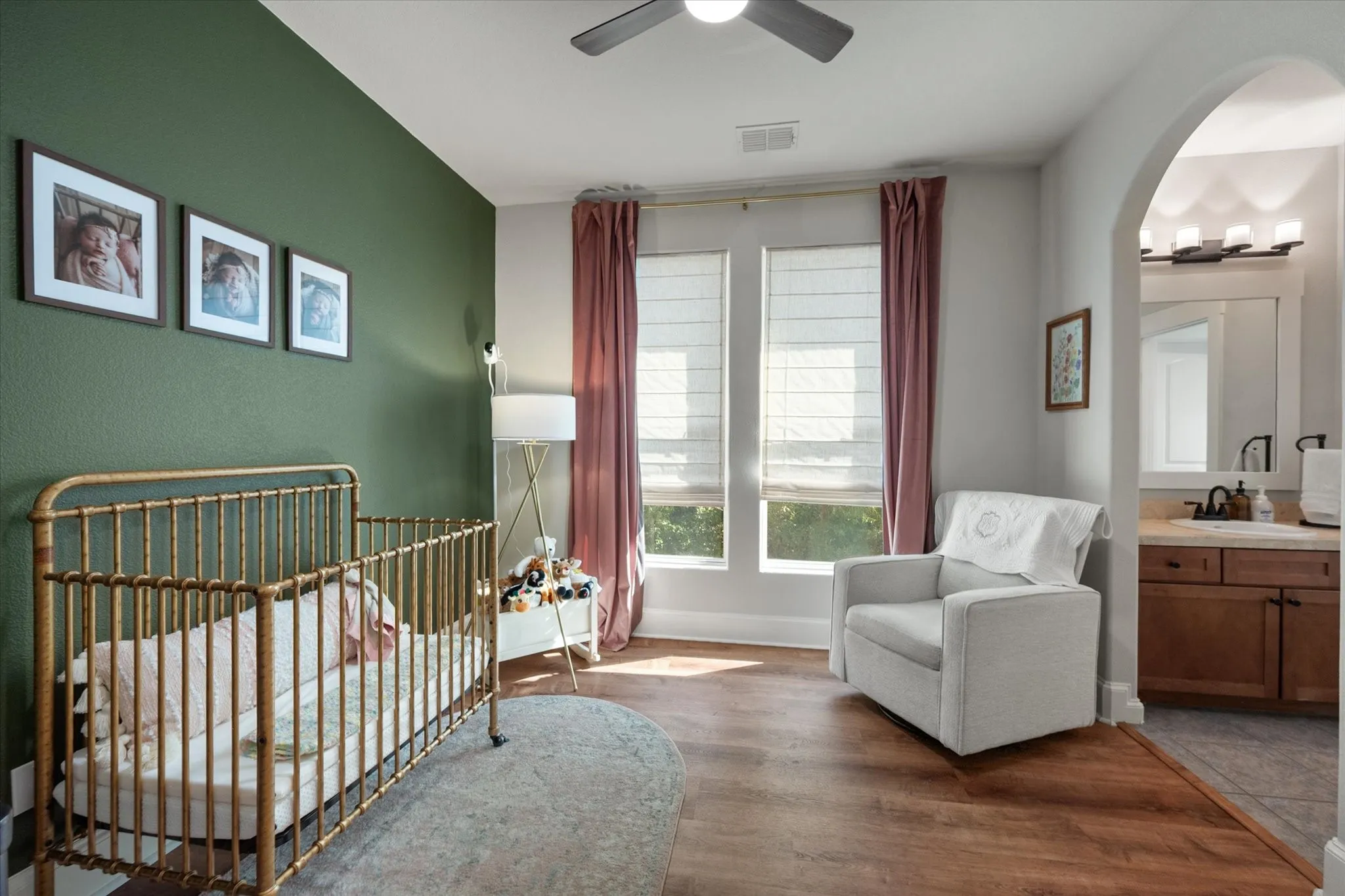 Wood-finish flooring, dual windows with Roman shades, a ceiling fan, and an arched doorway leading to a built-in vanity with a mirror and multi-light fixture