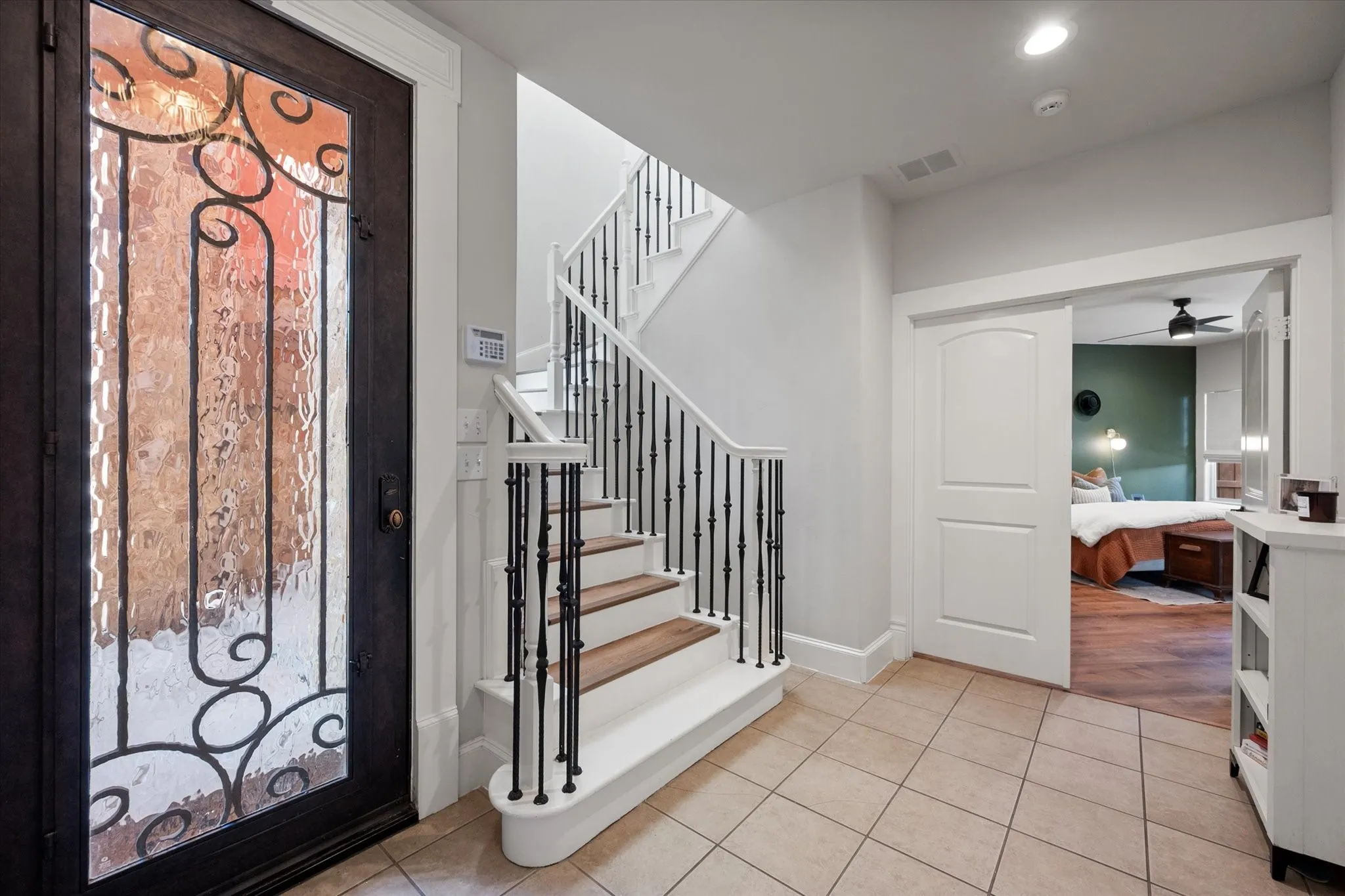 Foyer featuring front door with decorative glass and ironwork, tiled flooring, a wooden staircase with wrought iron balusters, wooden handrail and wood-finish treads and view of Primary Suite