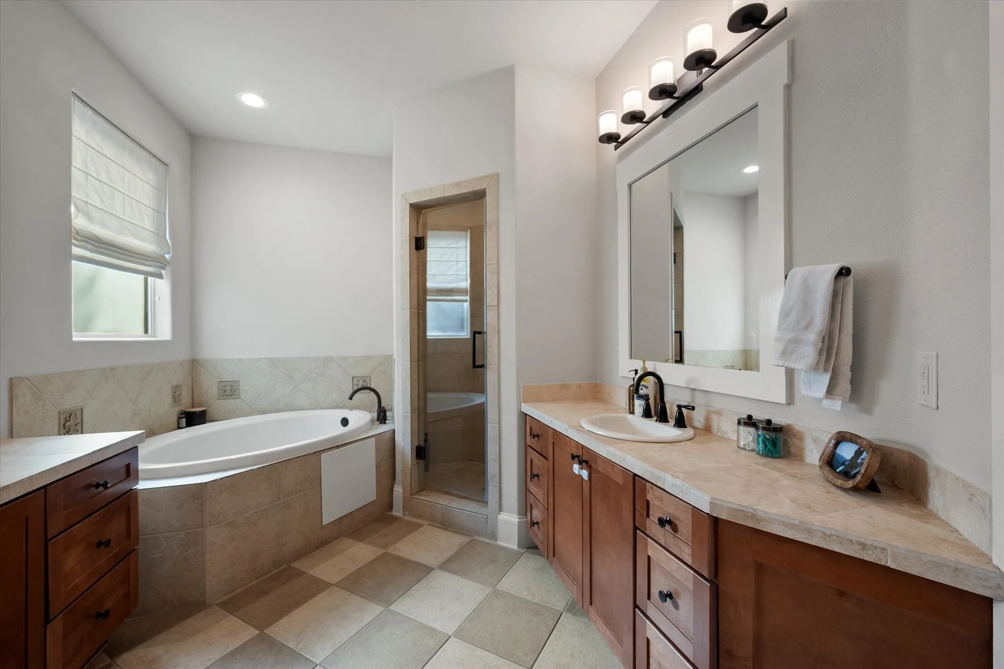 Corner soaking tub with tile surround, glass-enclosed shower, wood-finish vanity with countertop, and framed mirror with integrated lighting