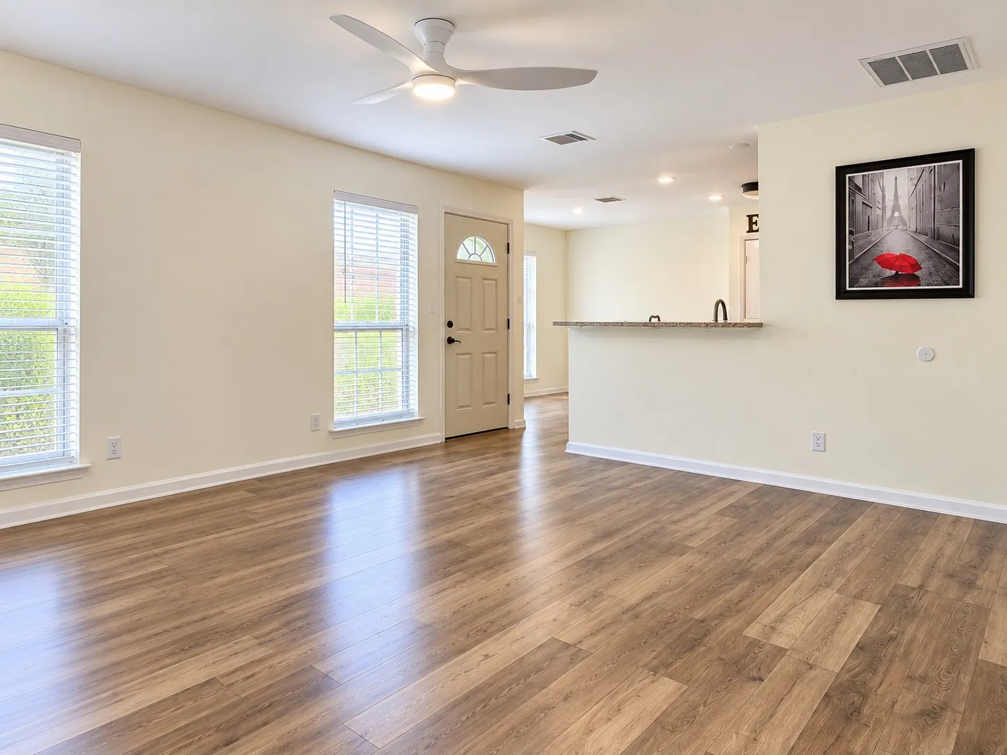 Wood-finish flooring, two large windows with blinds, a ceiling fan with light, and a breakfast bar with a stone countertop