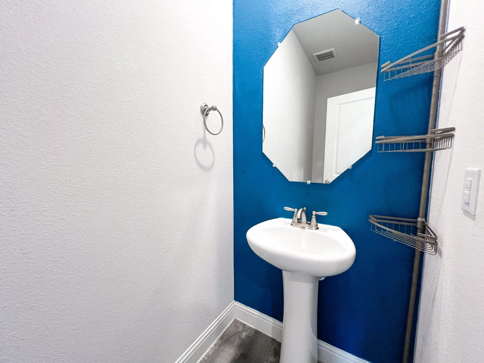 Pedestal sink with dual-handle faucet, octagonal mirror, corner shelving unit, and wood-finish flooring