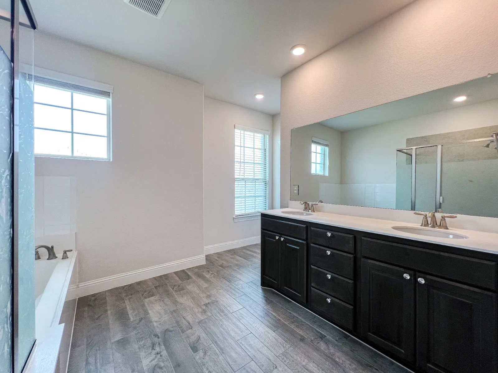 Spacious bathroom featuring wood-finish flooring, a double vanity with dark cabinetry and light countertops, a large wall-mounted mirror, a built-in bathtub, and a glass-enclosed shower