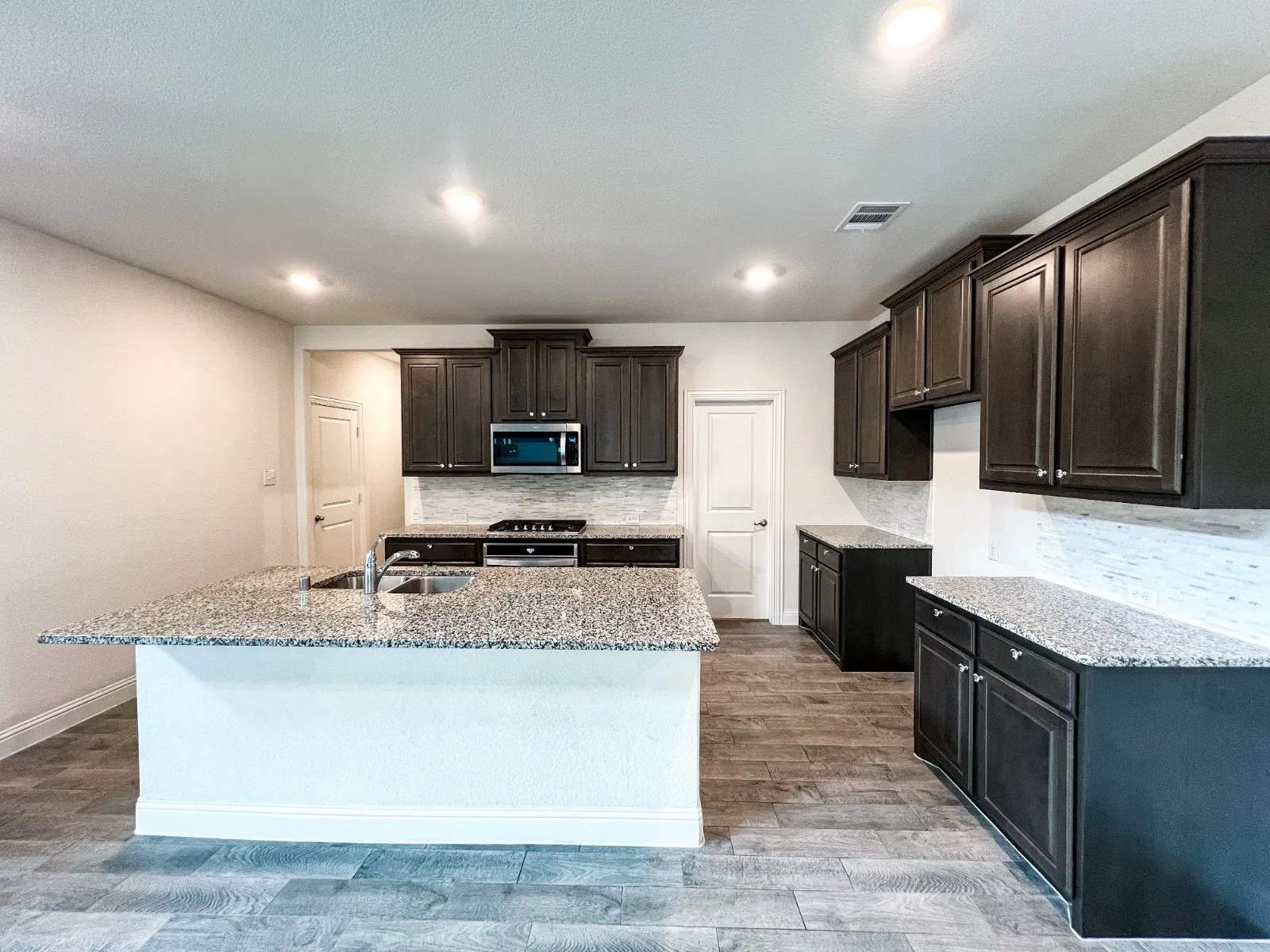 Kitchen featuring a central island with a sink, granite countertops, dark wood-finish cabinetry, a tile backsplash, and wood-look flooring