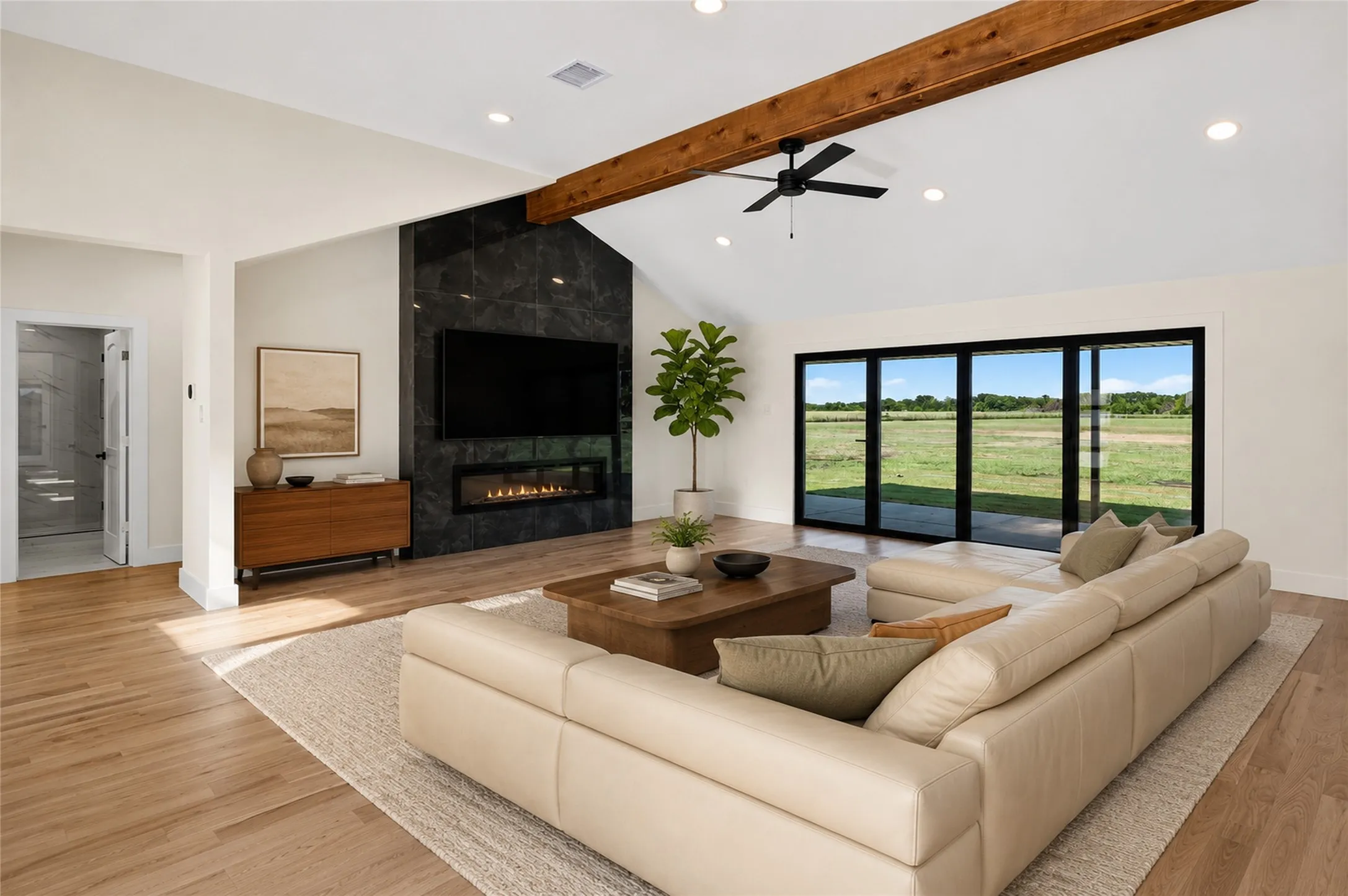 Spacious living area featuring a vaulted ceiling with an exposed wood beam, a black marble-style fireplace, and wide plank wood-finish flooring - Virtual Staging