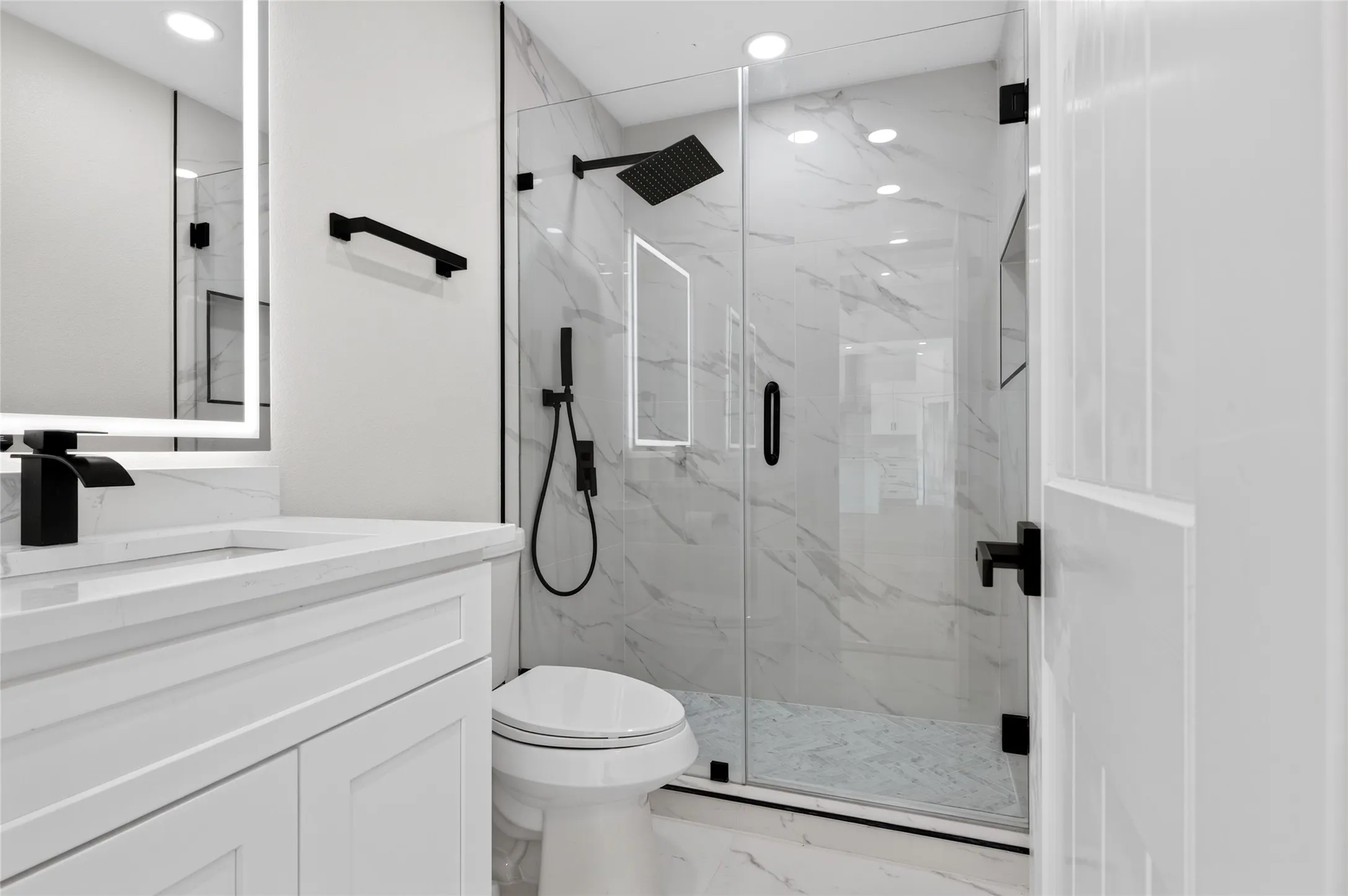 Bathroom featuring a walk-in shower with clear glass enclosure and black hardware, white vanity with quartz countertop, illuminated mirror, and black faucet