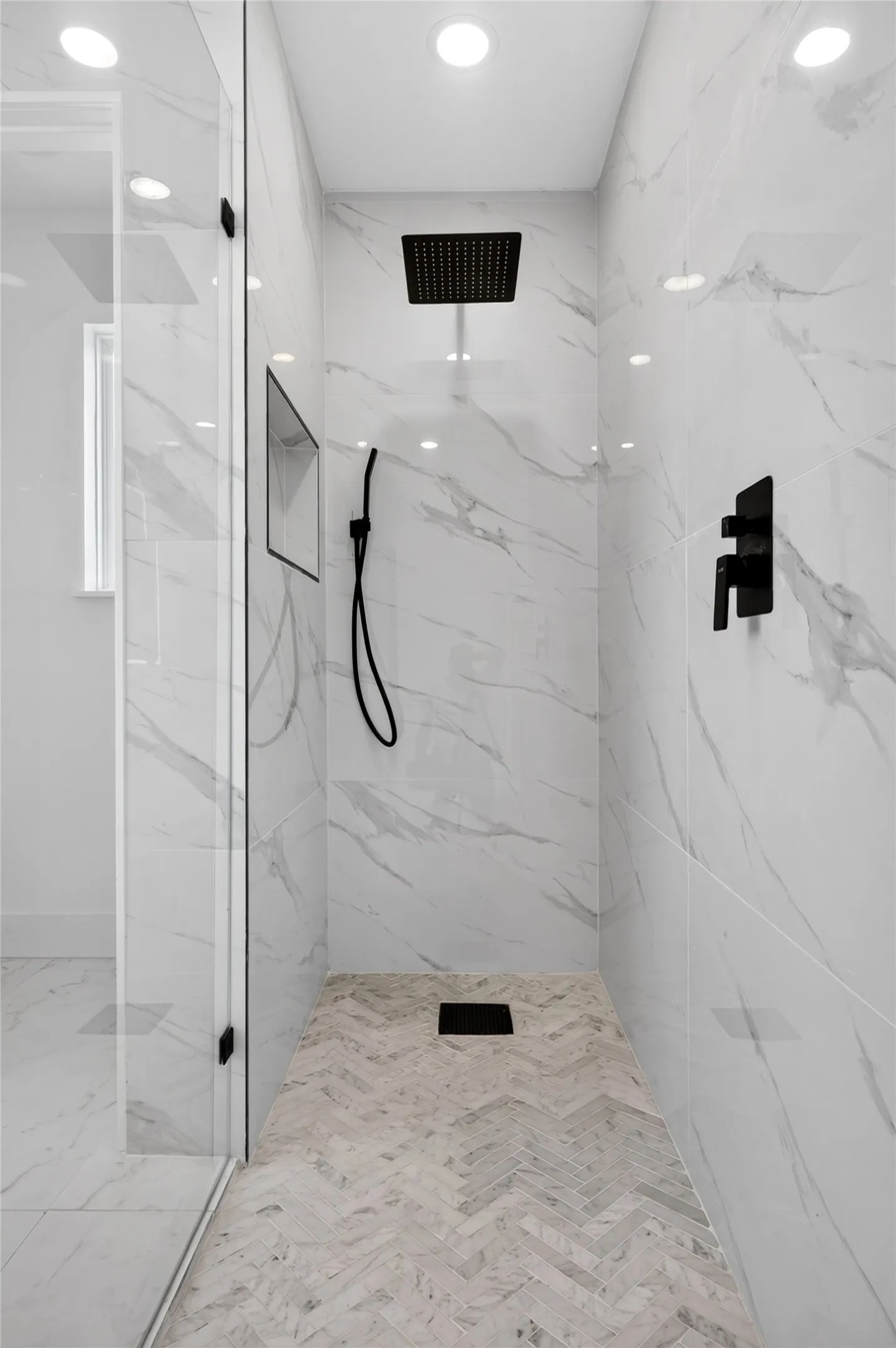Spacious shower enclosure featuring full-height marble-look tile walls, a herringbone-patterned floor, and frameless glass door