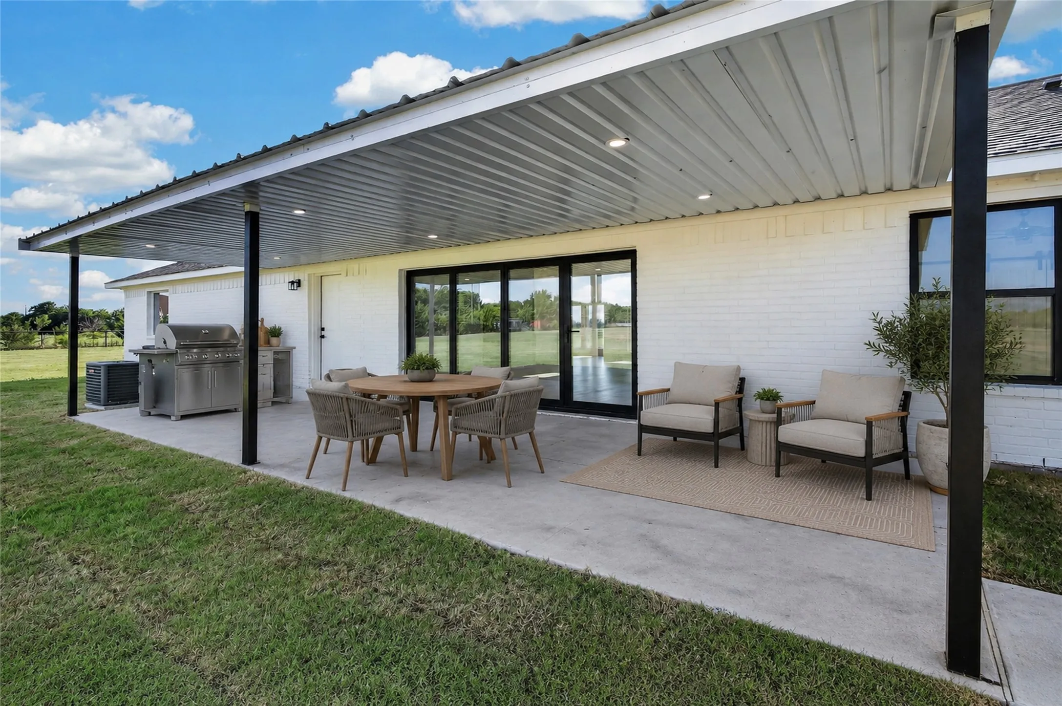 Covered patio featuring a corrugated metal roof with recessed lighting, a white brick exterior, black-framed windows, and expansive sliding glass doors - Virtually Staged