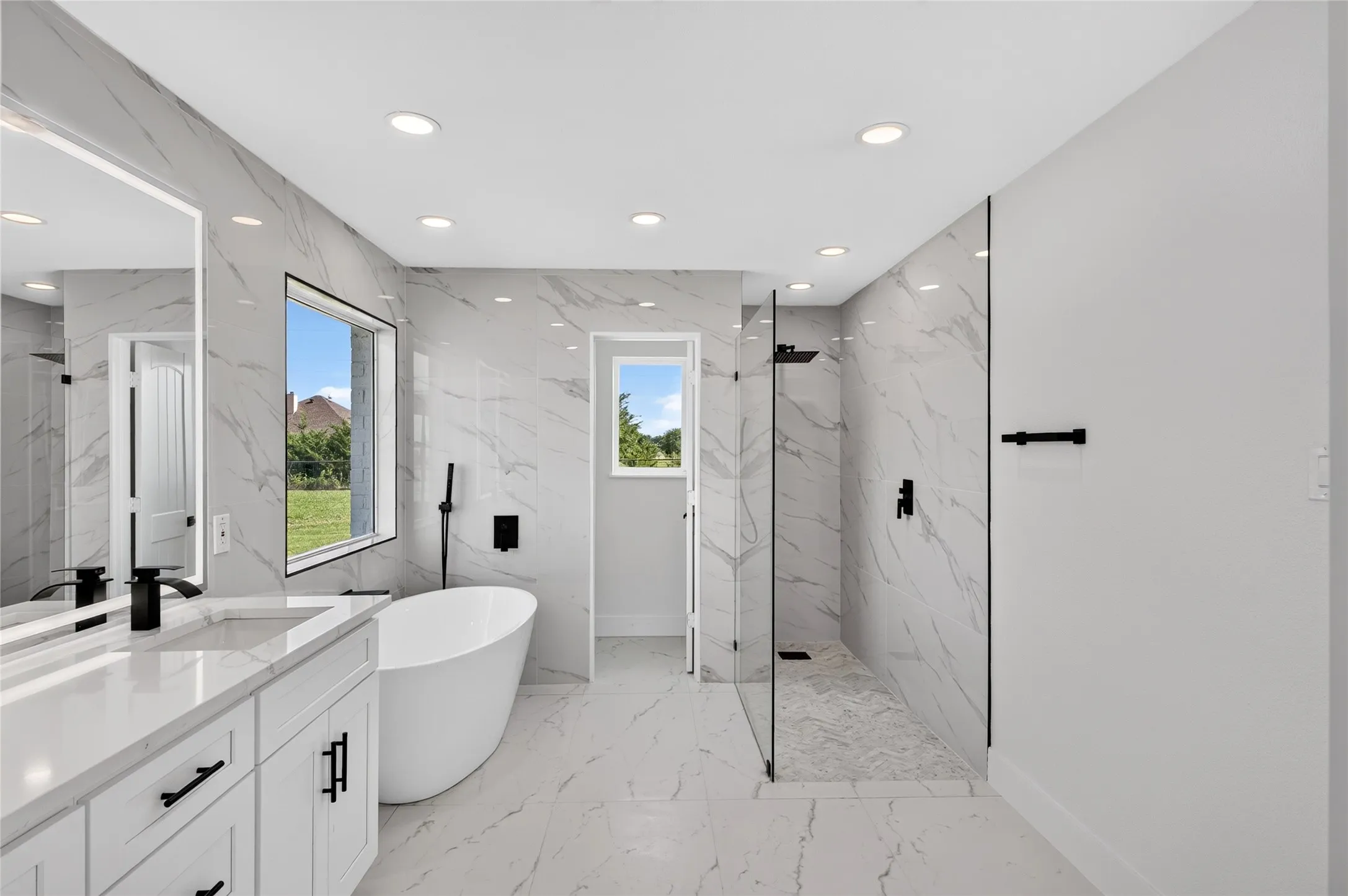 Modern bathroom featuring a freestanding soaking tub with a floor-mounted faucet, a spacious walk-in shower with glass partitions, a dual vanity with a white countertop and undermount sinks, and large-format tile flooring and wall accents