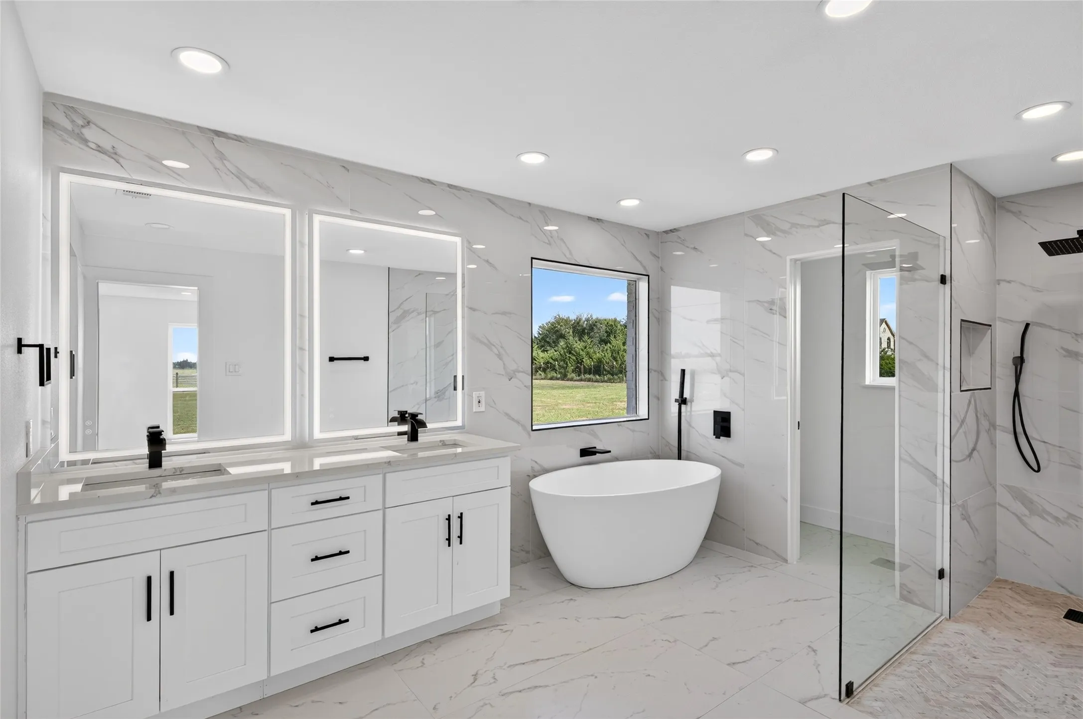 Bright bathroom featuring a dual vanity with white cabinetry and light-toned countertops, two illuminated mirrors, a freestanding soaking tub, and a spacious glass-enclosed shower with contrasting dark fixtures
