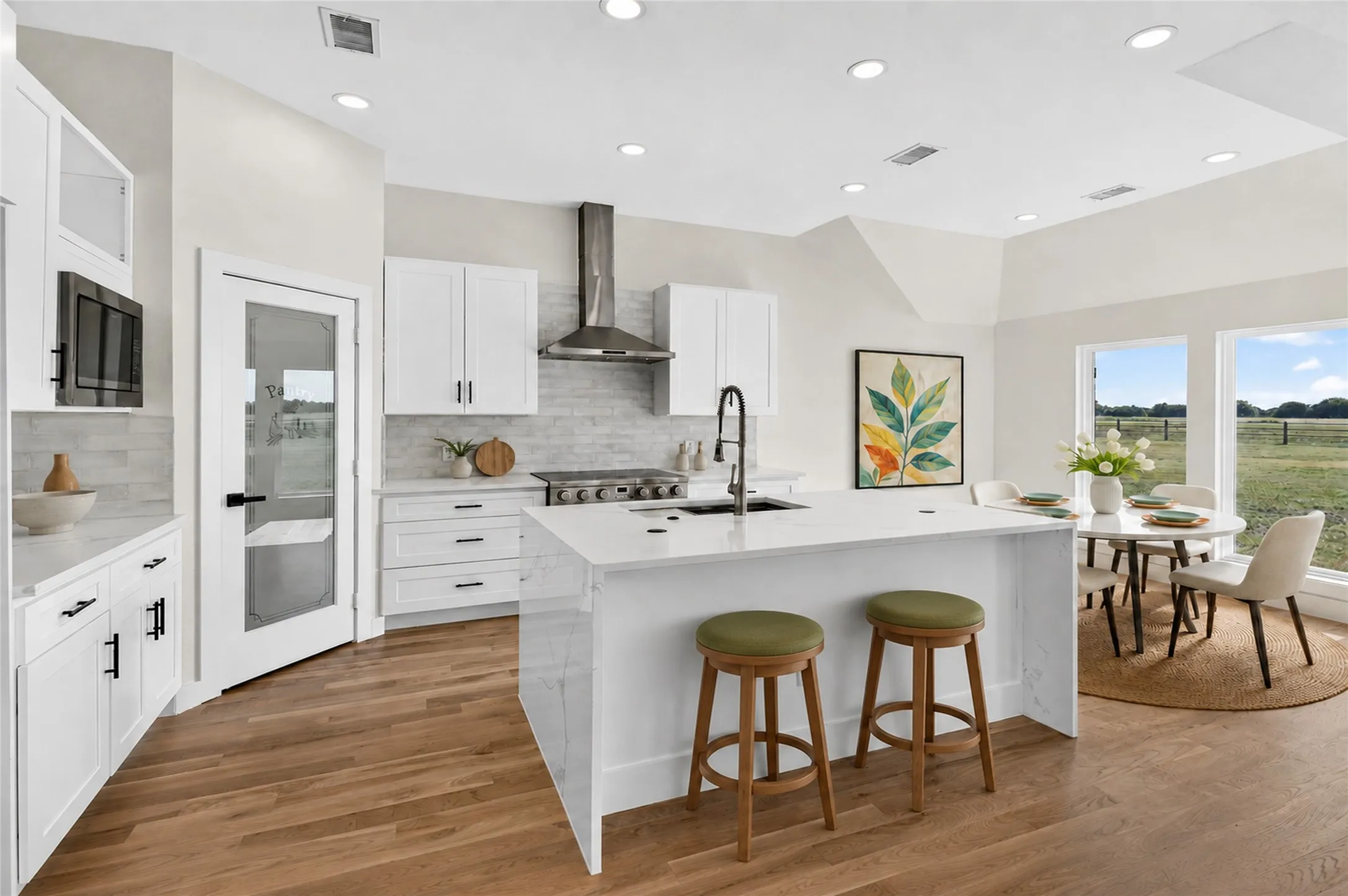 Modern kitchen featuring white cabinetry, a large center island with a sink, stainless steel range hood, wood-finish flooring, and a dining area with multiple windows - Virtually Staged