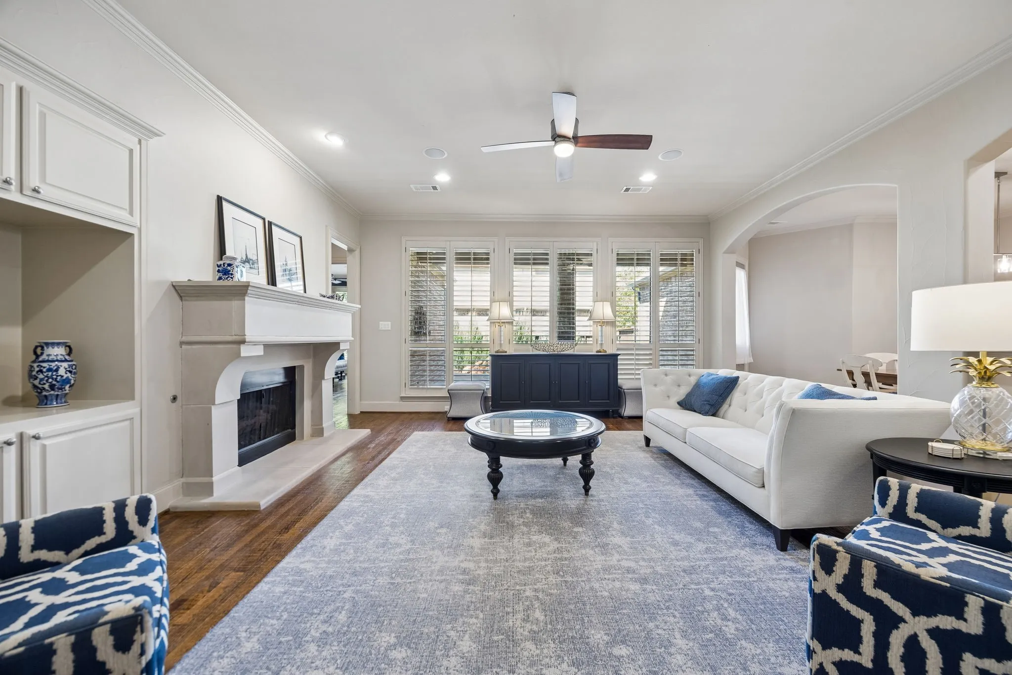 Main living area with a stone fireplace, built-in cabinetry, hand-scraped wood flooring, a ceiling fan, and plantation shutters
