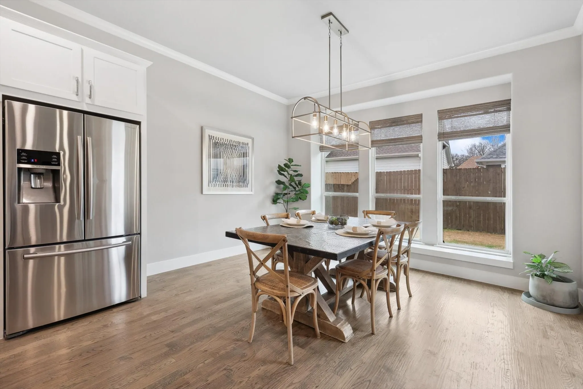 Stainless steel refrigerator, white shaker-style cabinetry, wood-finish flooring, contemporary linear chandelier, and triple windows with blinds