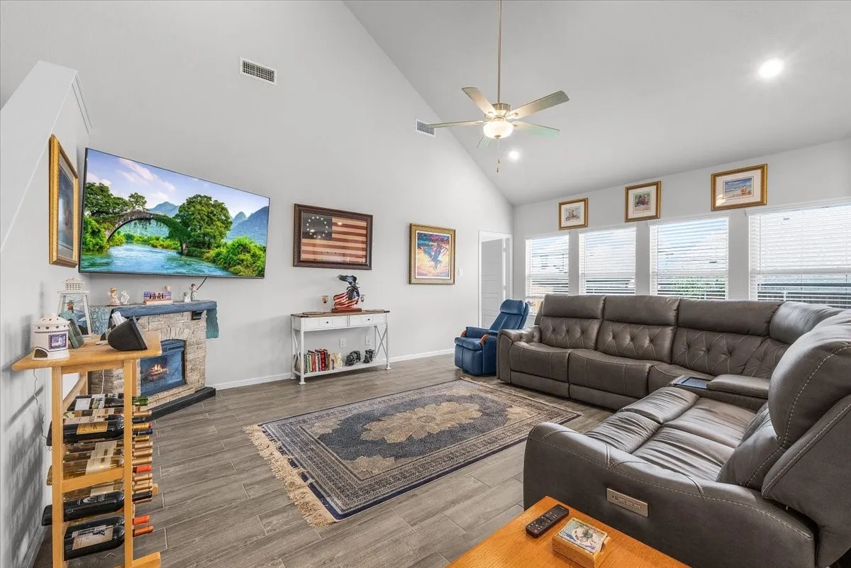 Vaulted ceiling, wood-finish flooring, stone-clad fireplace, ceiling fan with integrated lighting, and multiple windows