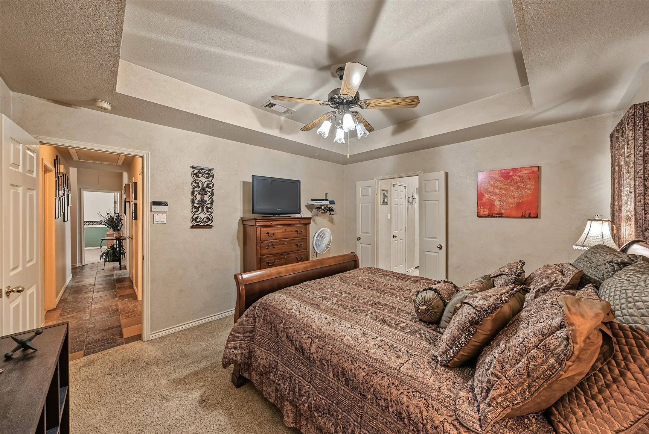 Tray ceiling with a ceiling fan, carpet flooring, and a doorway to a tiled hallway