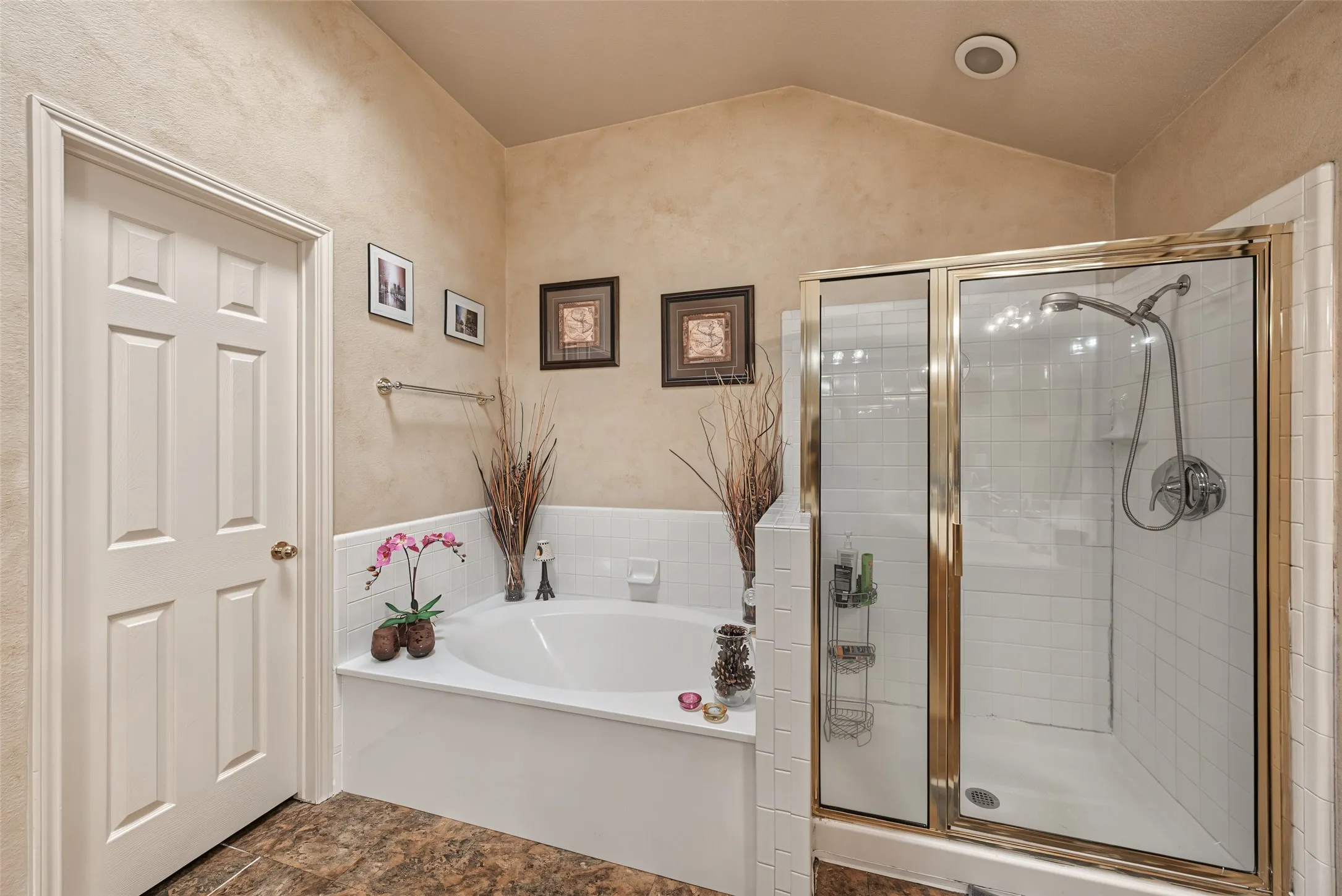 Built-in tub with white tile surround, glass-enclosed shower with brass-finish framing, vaulted ceiling, and panel door