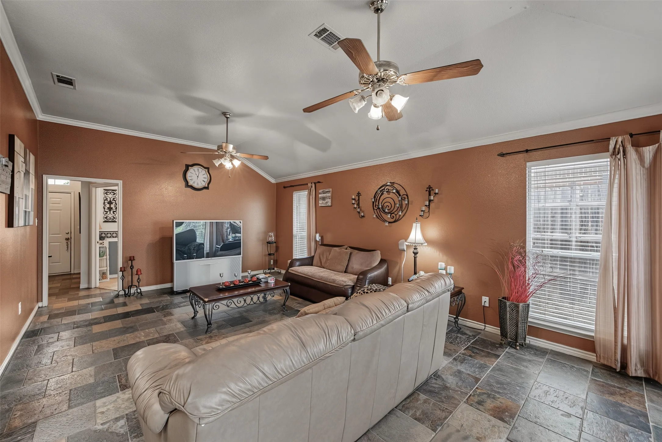 Vaulted ceiling, crown molding, two ceiling fans with lighting, slate-style flooring, and a large window with blinds