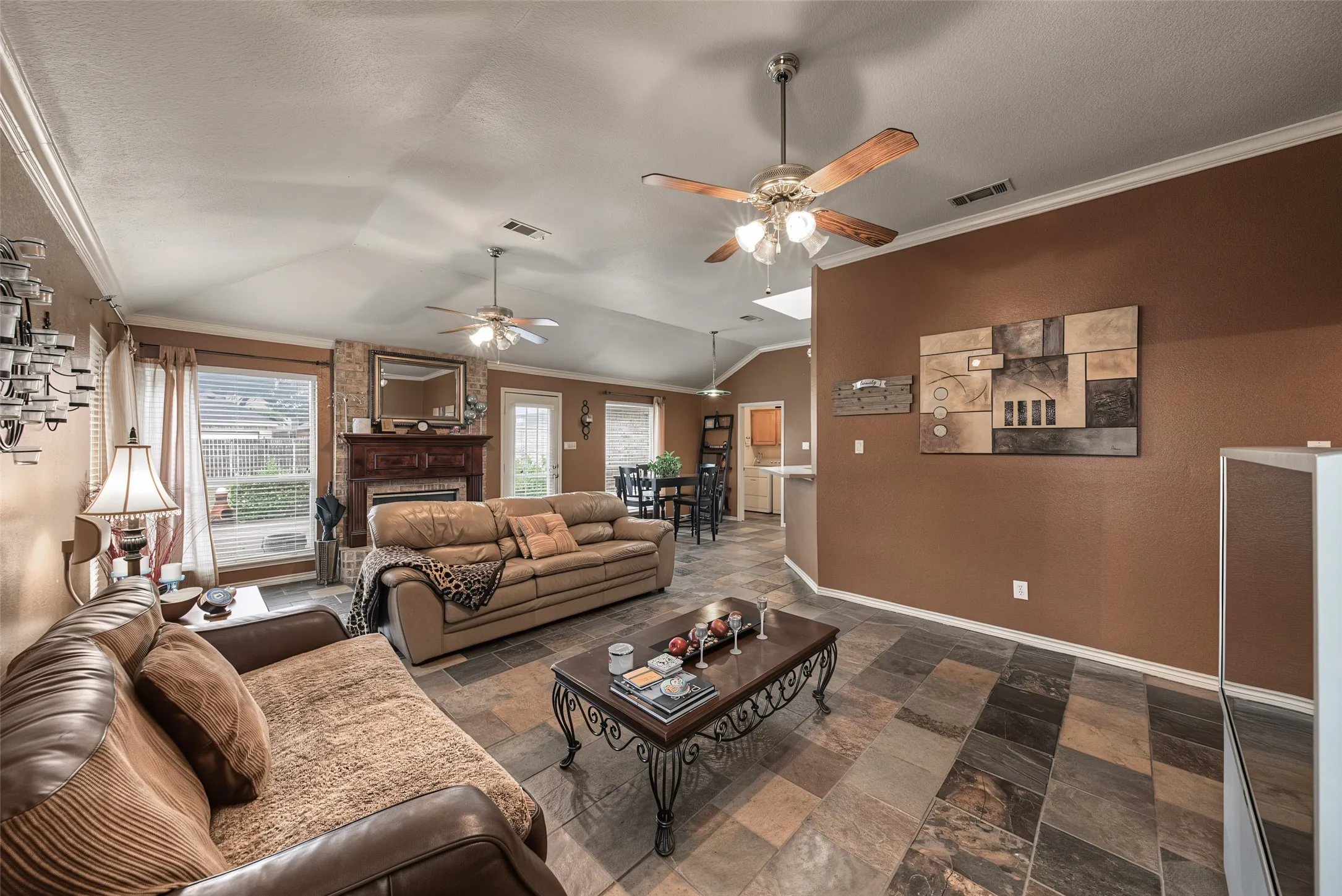 Vaulted ceiling, stone-clad fireplace with wood mantel, large windows, ceiling fans, and multi-tone tile flooring